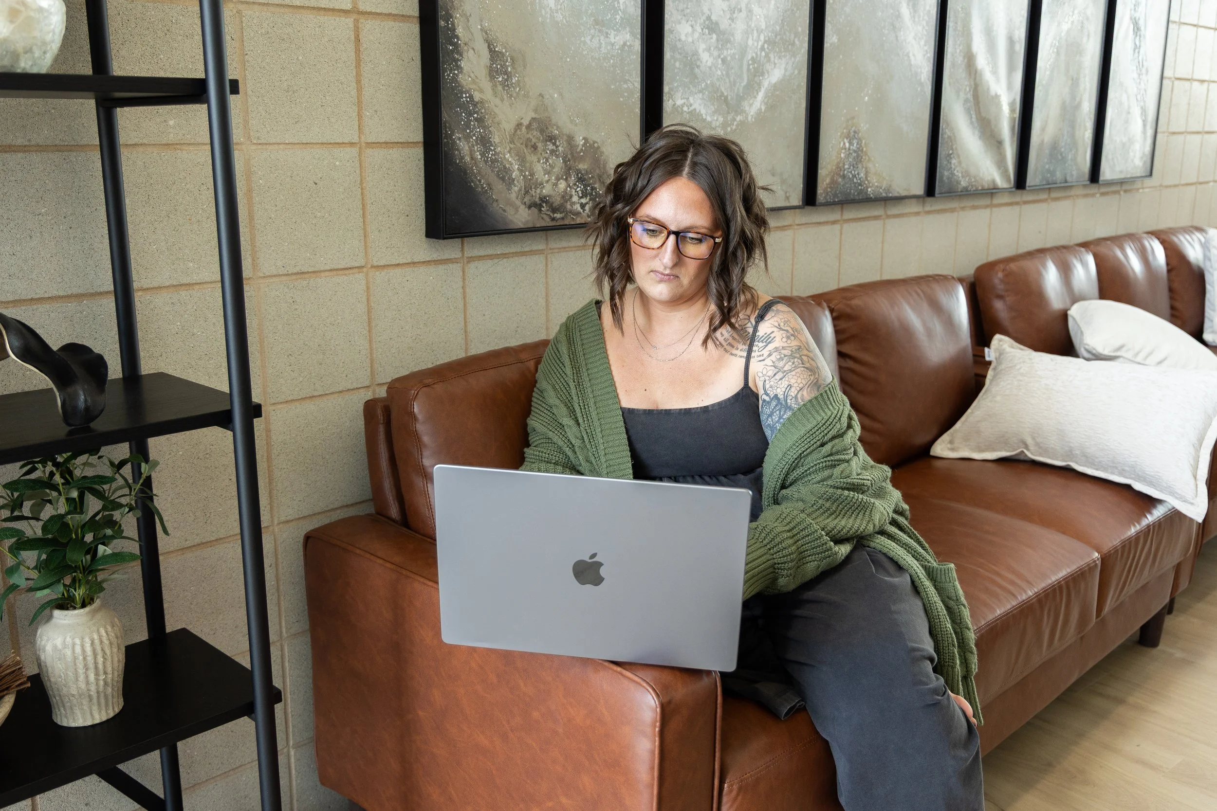 A woman with glasses and dark hair sitting on a brown leather couch working on a silver laptop. She is wearing a green cardigan and black clothing, with decorative tattoos on her right arm. Behind her, there's abstract artwork on a beige brick wall, and to her left, a black shelf with a ceramic vase, a plant, and a black sculpture.