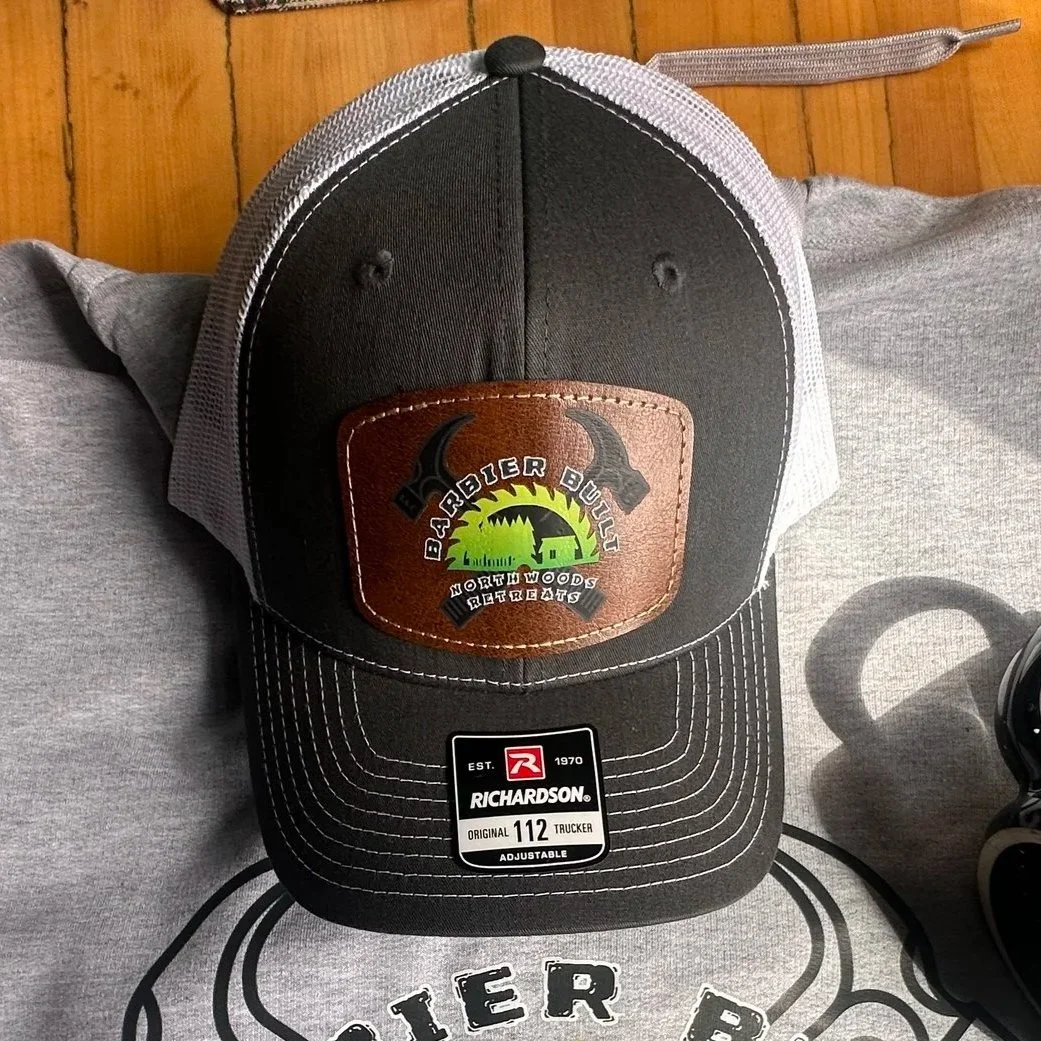 A black and gray baseball cap with a leather patch on the front. The patch has two horns and a sunset with trees and a house, along with the words "Barbier Buzz" and "North Woods Retreats." The cap has white stitching and is placed on a gray surface with a drawing of a mountain and text partially visible.