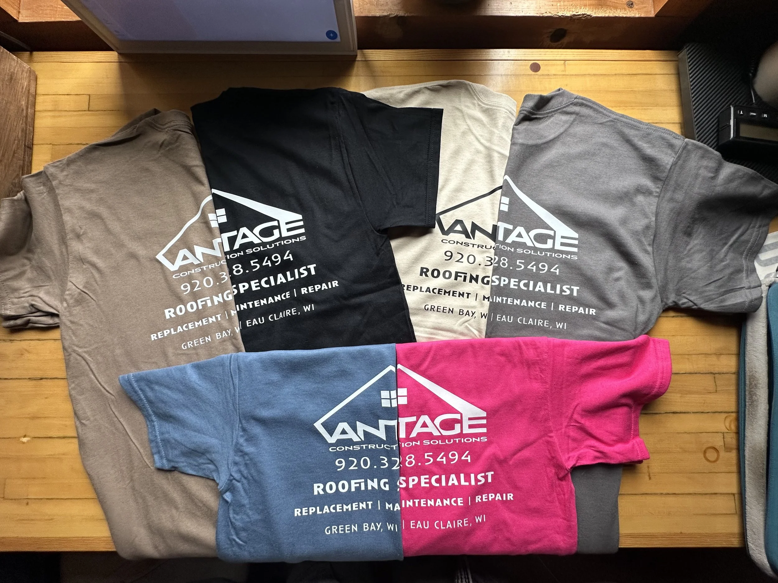 Four folded T-shirts with a company logo and contact information printed on them, laid out on a wooden surface. The shirts are in beige, black, gray, and pink colors.
