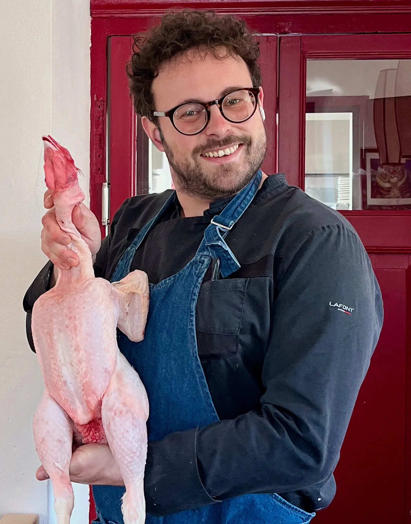 A man with glasses and a beard, smiling, holds a raw, plucked chicken. He is wearing a black shirt and a blue apron. The background shows a red door and part of a kitchen.