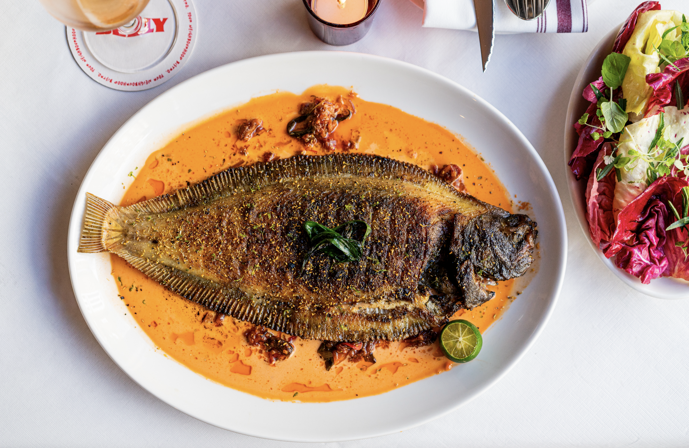 A whole cooked fish served on a white plate with orange sauce, garnished with herbs and a lime wedge. A side salad with mixed greens and colorful lettuce is in a bowl to the right. Part of a glass of beer and a drink are visible at the top.