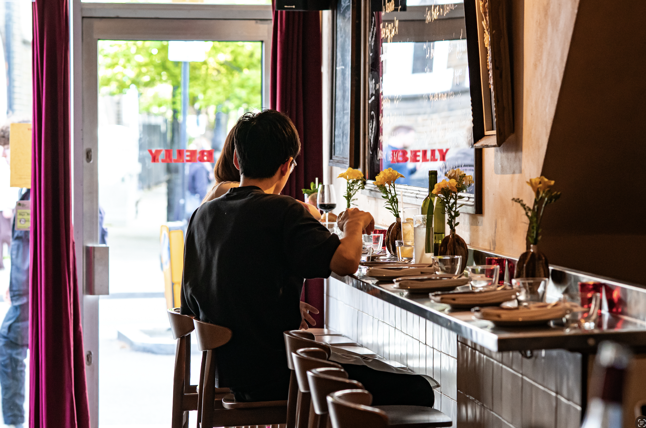 Inside a cozy restaurant with a bar counter, where a man is sitting with his back to the camera, having a glass of red wine. The restaurant has large windows with greenery visible outside, red curtains, and small vases with flowers on the counter.
