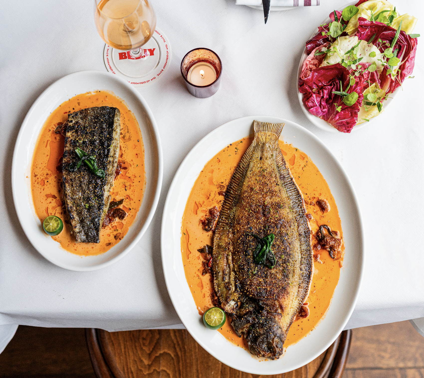 Two plates of cooked fish served with an orange-colored sauce, garnished with chopped herbs and lime slices, on a white tablecloth. A glass of rosé wine, a lit candle, and a salad with radicchio, lettuce, and fresh greens are also on the table.