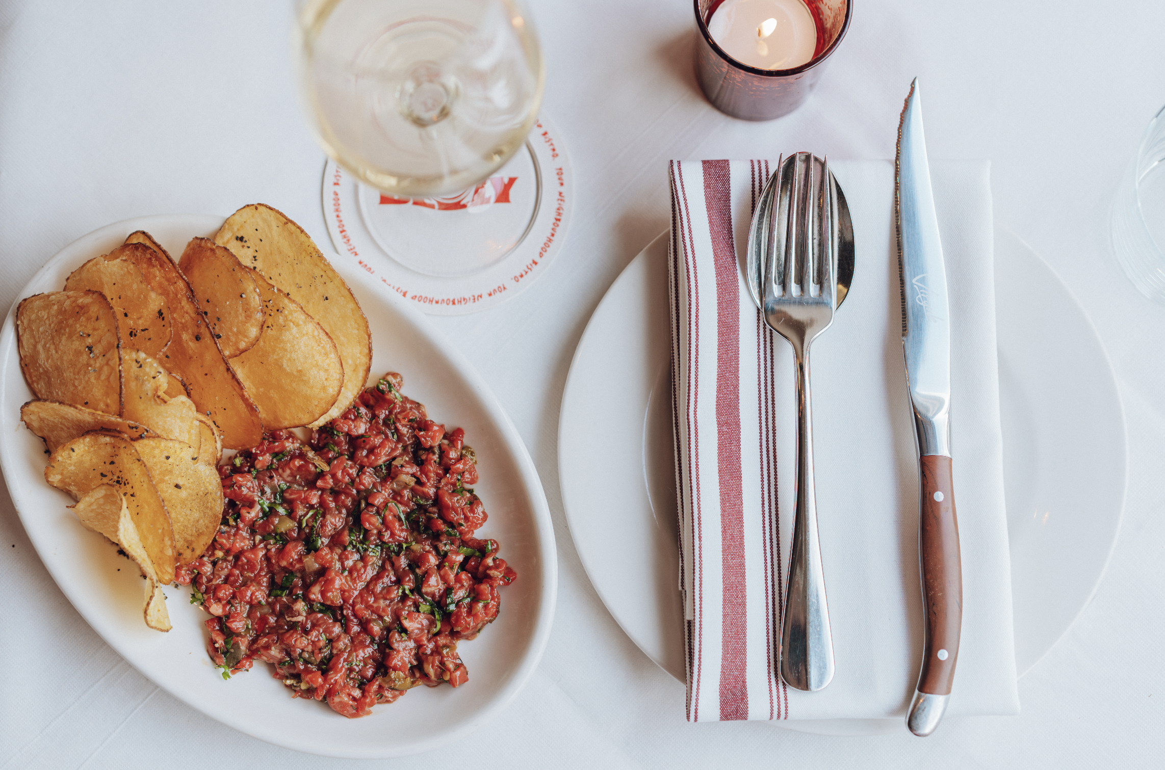 A white plate with potato chips and a serving of steak tartare, set on a white table with a glass of white wine, a lit candle in a red holder, and a white napkin with silverware in a restaurant setting.