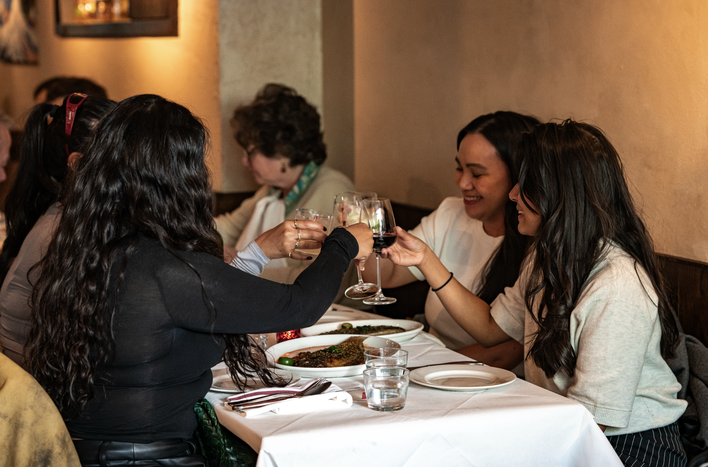 People at a restaurant table clinking wine glasses, smiling, with food on the table and warm lighting.