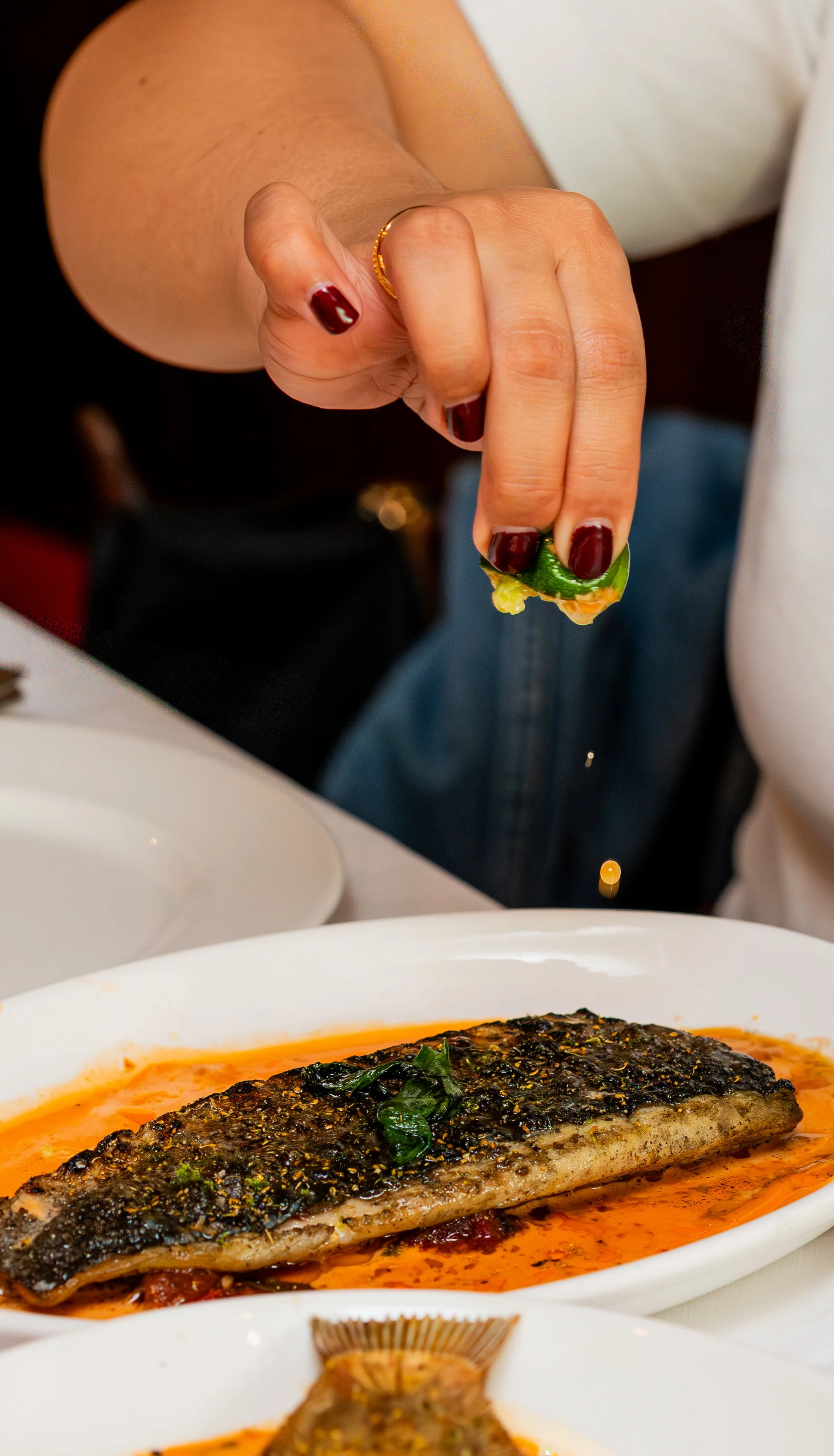 Close-up of a person sprinkling herbs or seasoning on a cooked fish fillet in a white dish, with other dishes partially visible in the background.