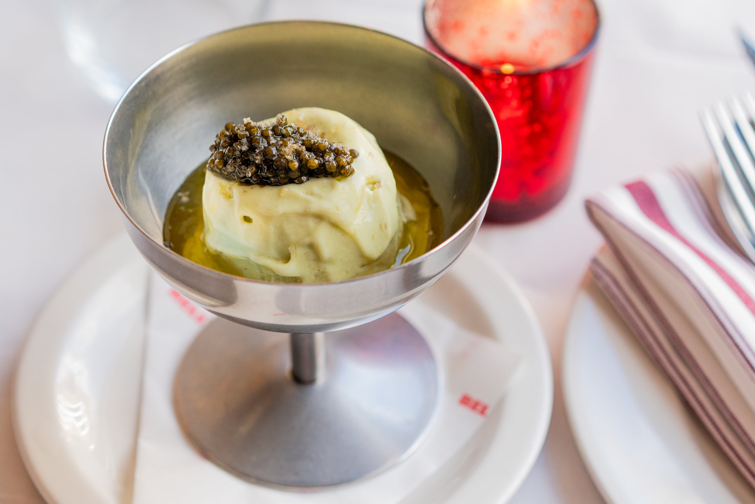 A dessert glass with a scoop of vanilla ice cream topped with black caviar, with a red drink in the background and utensils on a striped napkin to the side.