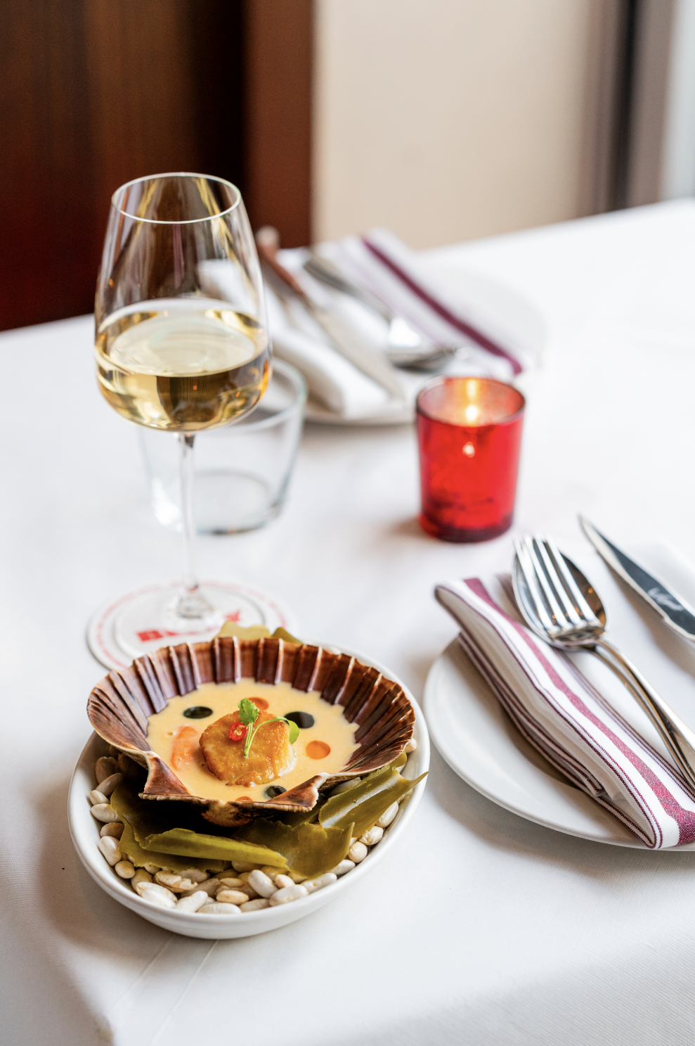 A glass of white wine on a table set with cutlery, a red candle holder, and a scallop shell with food served inside, garnished with herbs, surrounded by green leaves, on a white tablecloth.