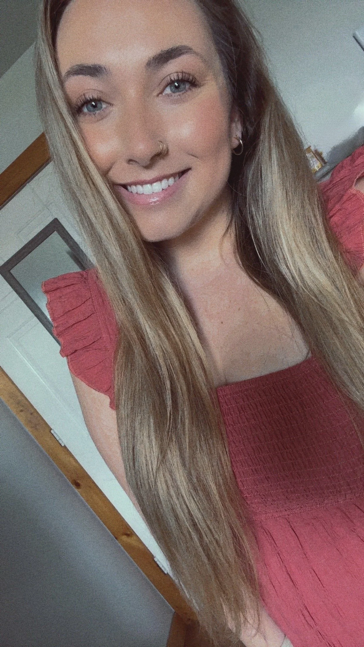 Close-up of a young woman with long light brown hair, blue eyes, and a bright smile, wearing a red top with ruffled sleeves, in a room with white walls and a door in the background.