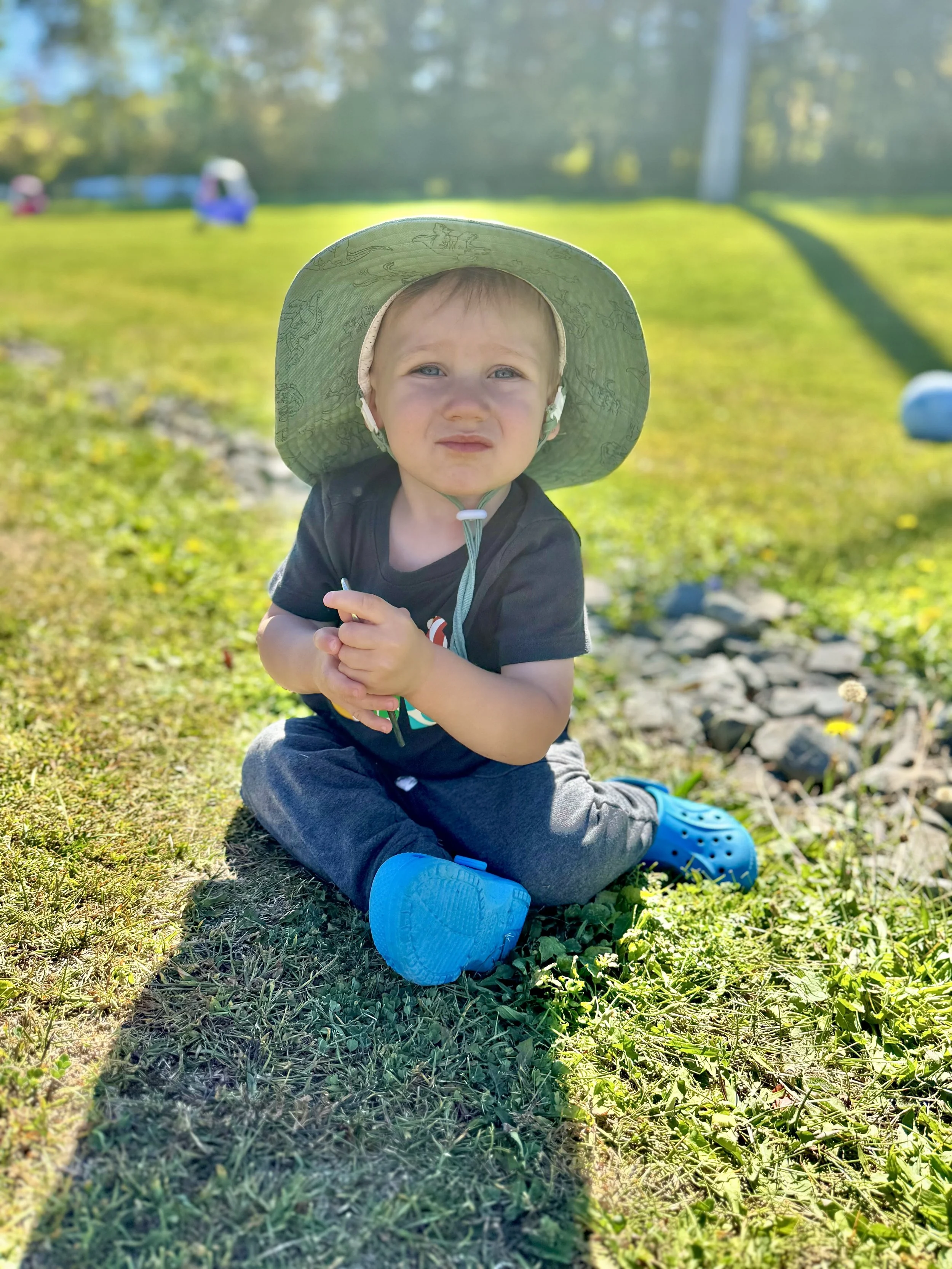 Young child sitting on grass outdoors, wearing a large sun hat, black t-shirt, grey pants, and bright blue crocs, with a curious expression, in bright sunlight with a blurred background of trees and a small blue toy.