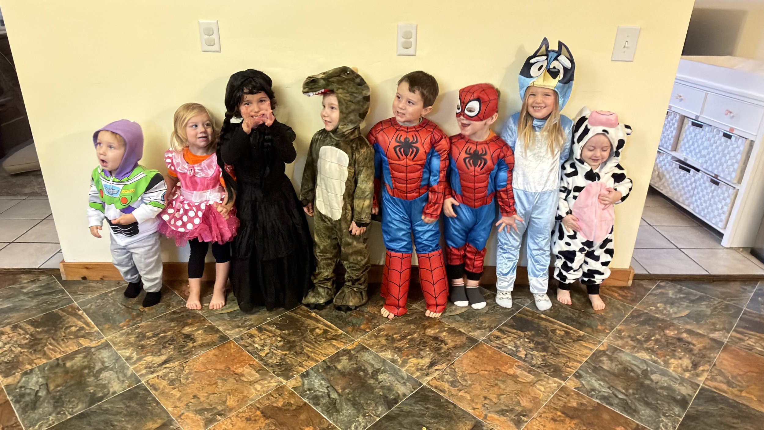 Children dressed in various costumes, including Buzz Lightyear, a princess, a black dress, a dinosaur, Spider-Man, a blue animal onesie, a cow, and a pink unicorn, standing against a yellow wall with electrical outlets.