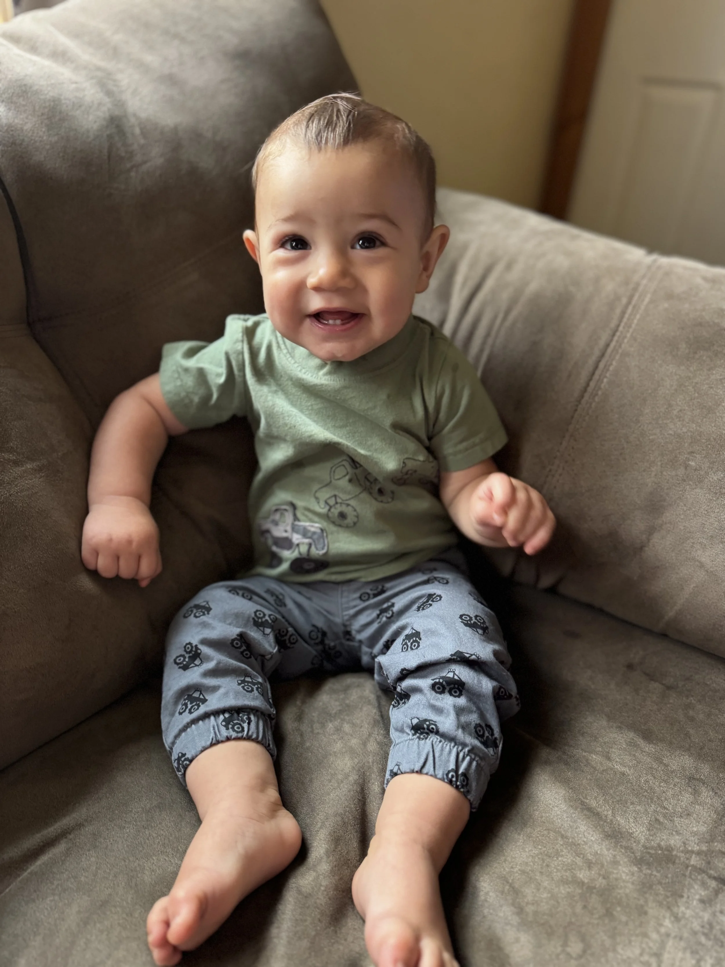 Smiling baby sitting on a beige couch, wearing a green T-shirt and gray pants with vehicle prints.
