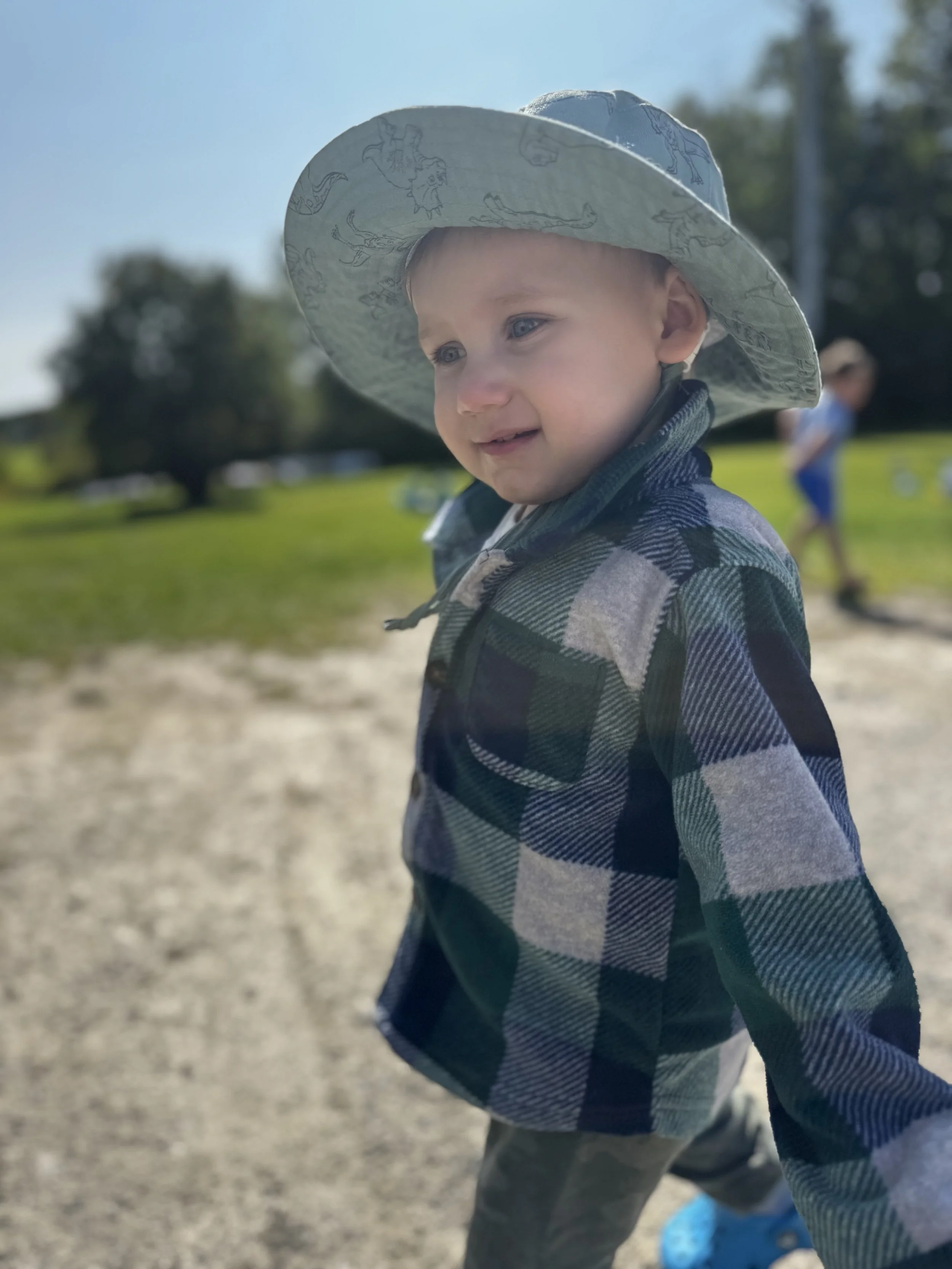 A young boy wearing a large sun hat and a plaid jacket smiling outdoors on a sunny day with trees and grass in the background.