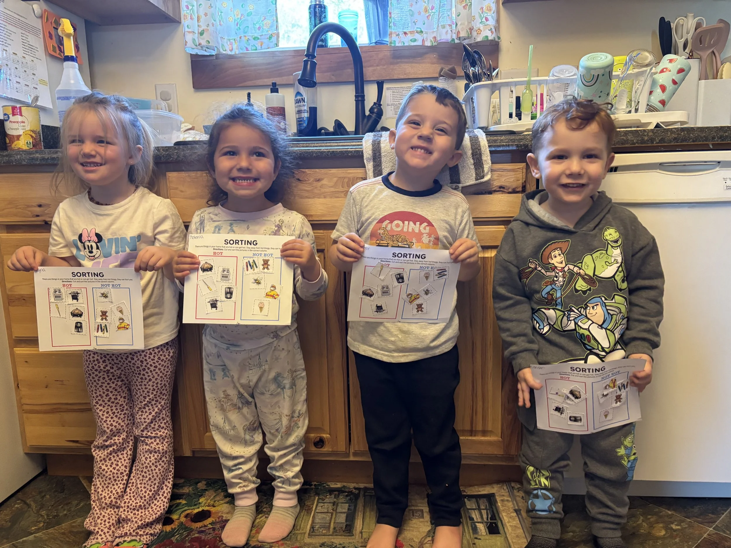 Four young children standing in a kitchen holding sorting worksheets. The children have happy expressions and are dressed in casual pajamas and t-shirts. The kitchen background features a counter with various kitchen items and a window with colorful 