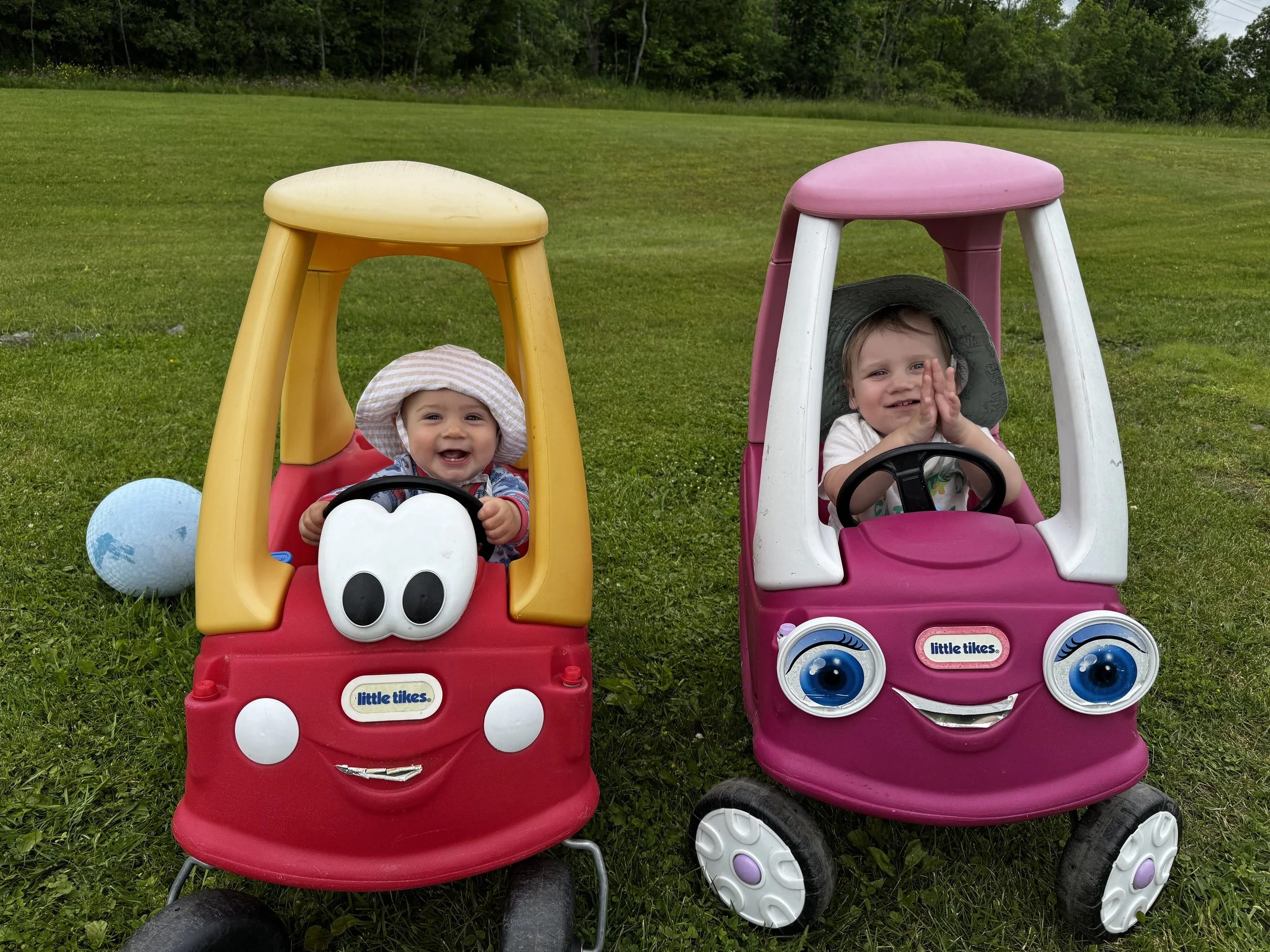 Two smiling children sitting in colorful toy cars outdoors on green grass, with trees in the background. The child on the left is in a red and yellow car with a face design, wearing a sunhat, while the child on the right is in a pink and white car wi