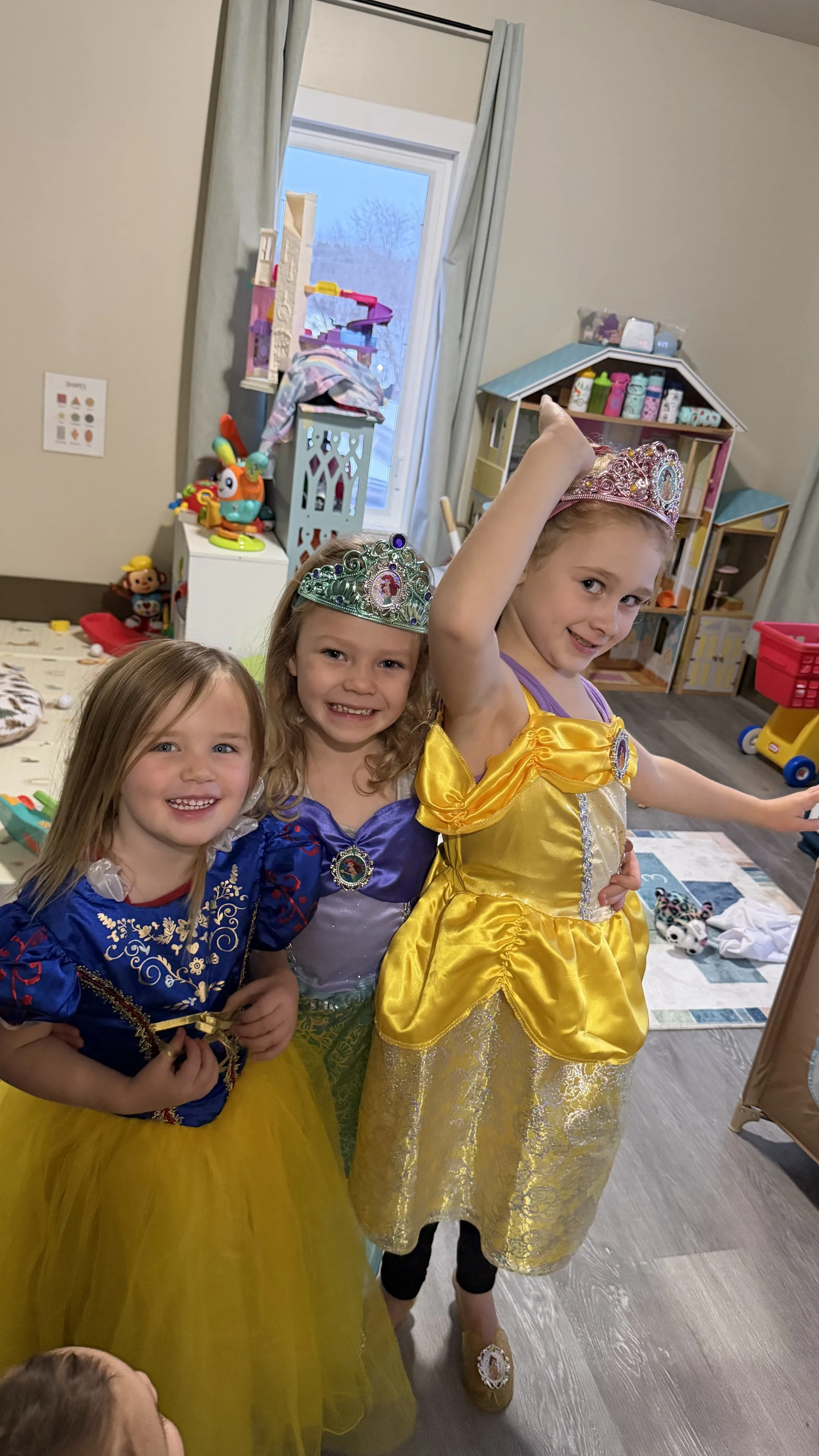 Three young girls dressed as princesses, wearing tiaras and colorful gowns, smiling and posing together in a playroom.