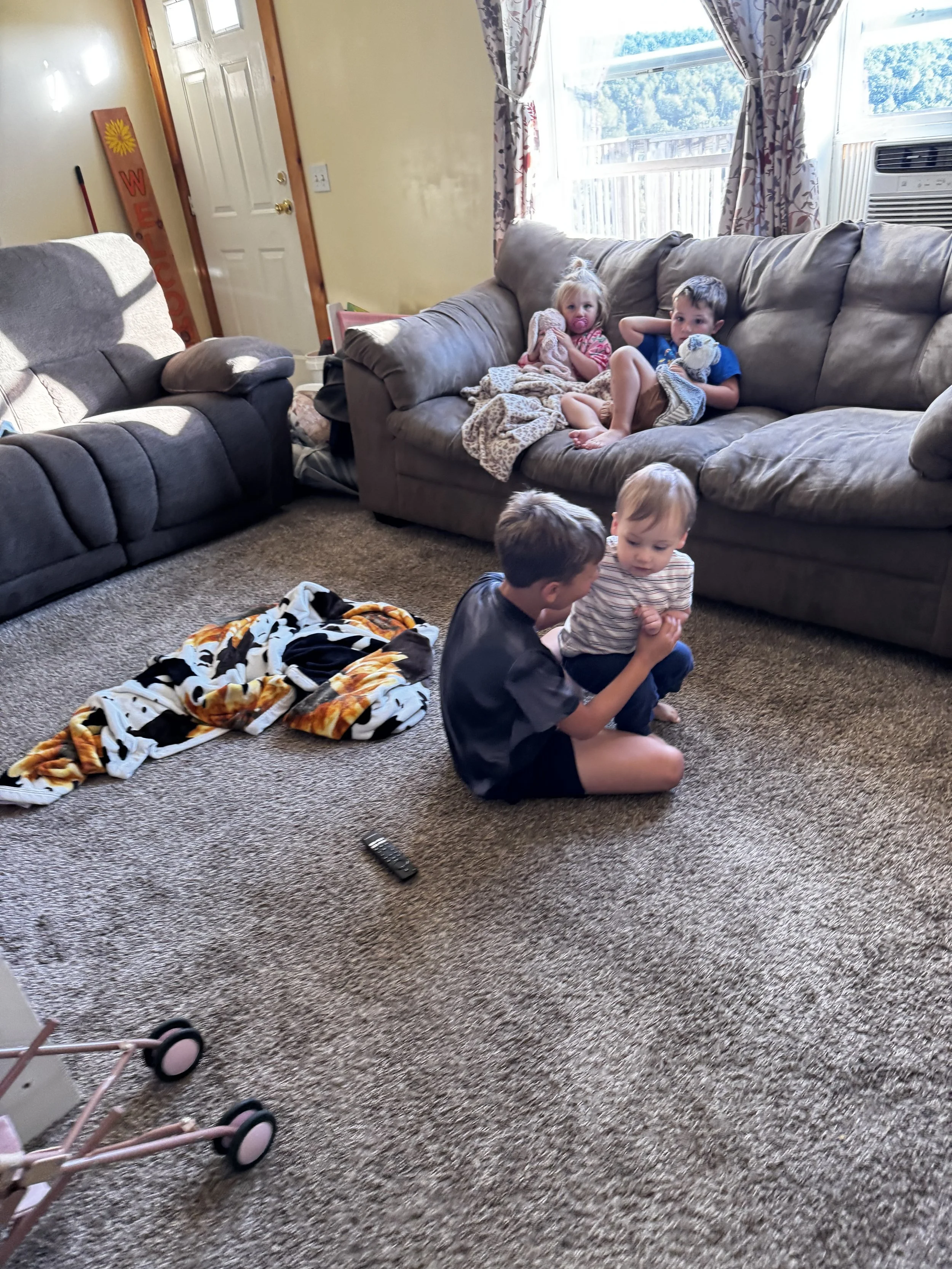 Four children in a living room with beige carpet and a large window. Two children are sitting on the floor facing each other, talking. One boy is holding a small girl on his lap. Two other children are sitting on a couch watching.