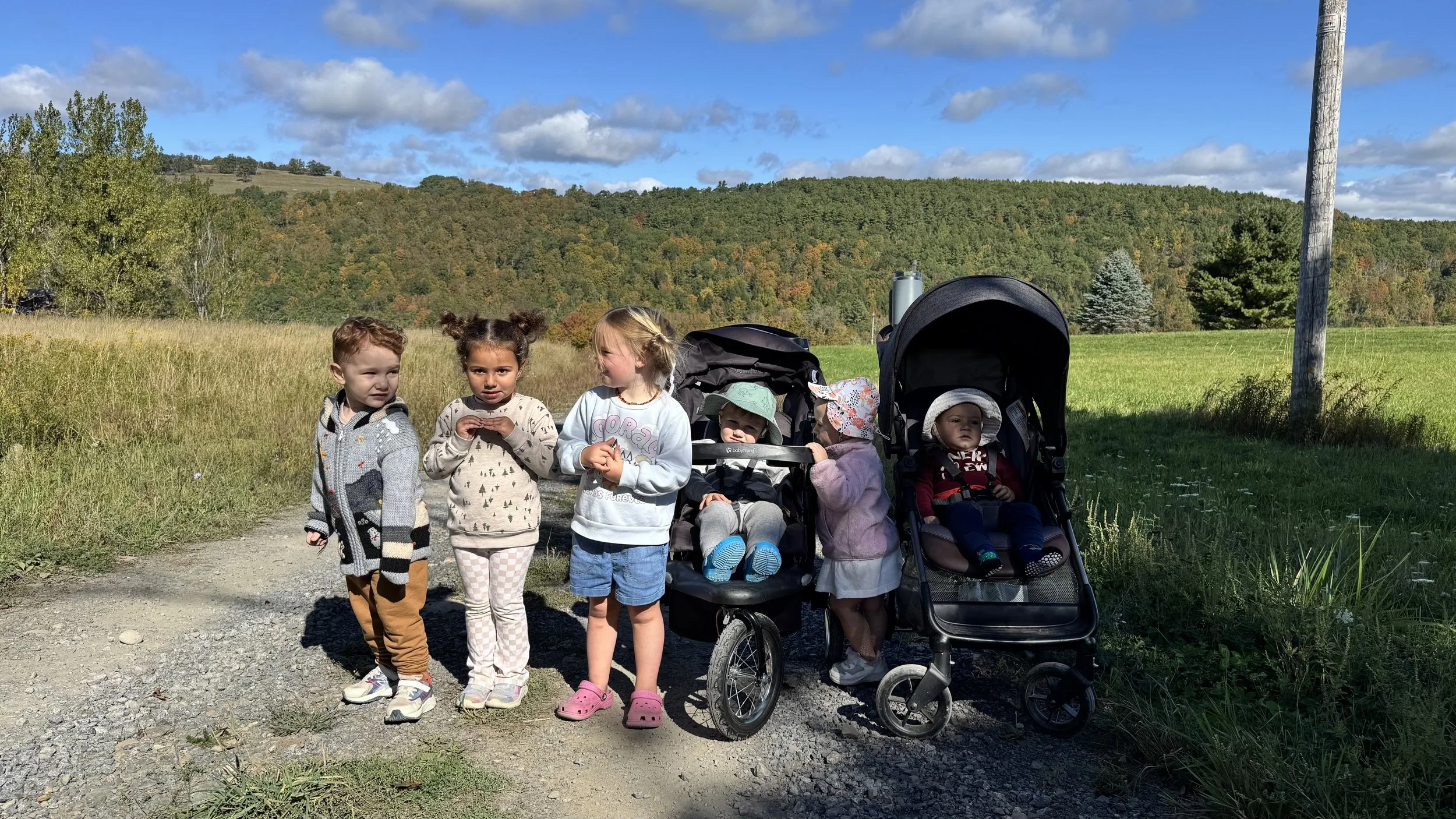 Group of five children standing on a dirt path in nature, with green grass, trees, and hills in the background under a partly cloudy sky.