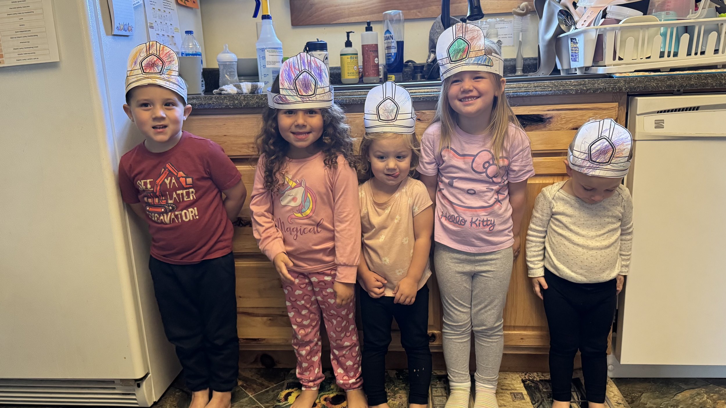 Five children stand in a kitchen wearing paper chef hats. They are smiling and facing the camera. The children are dressed in casual pajamas.