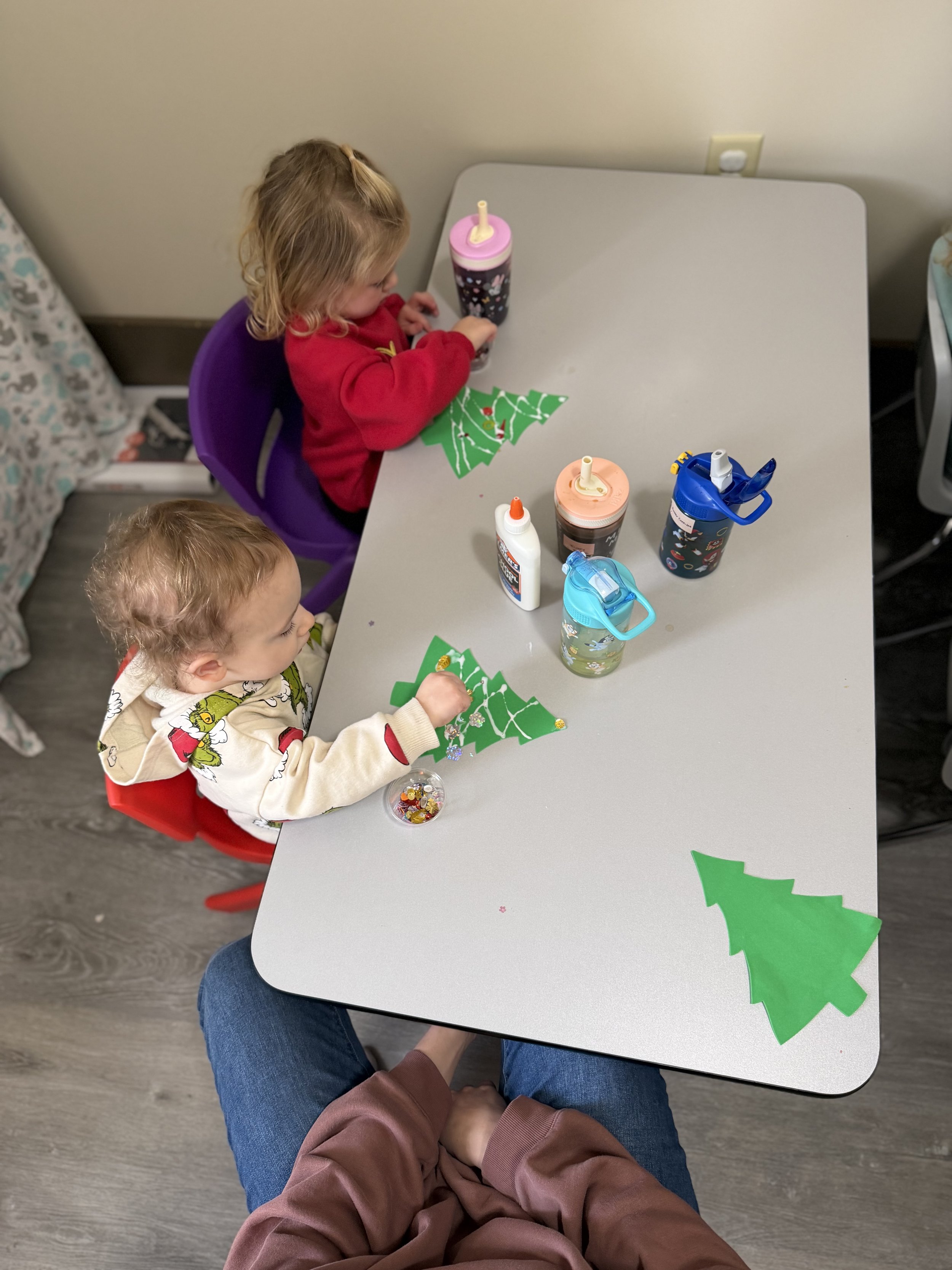 Two young children sitting at a table decorating paper Christmas trees with stickers and engaging in holiday craft activities.