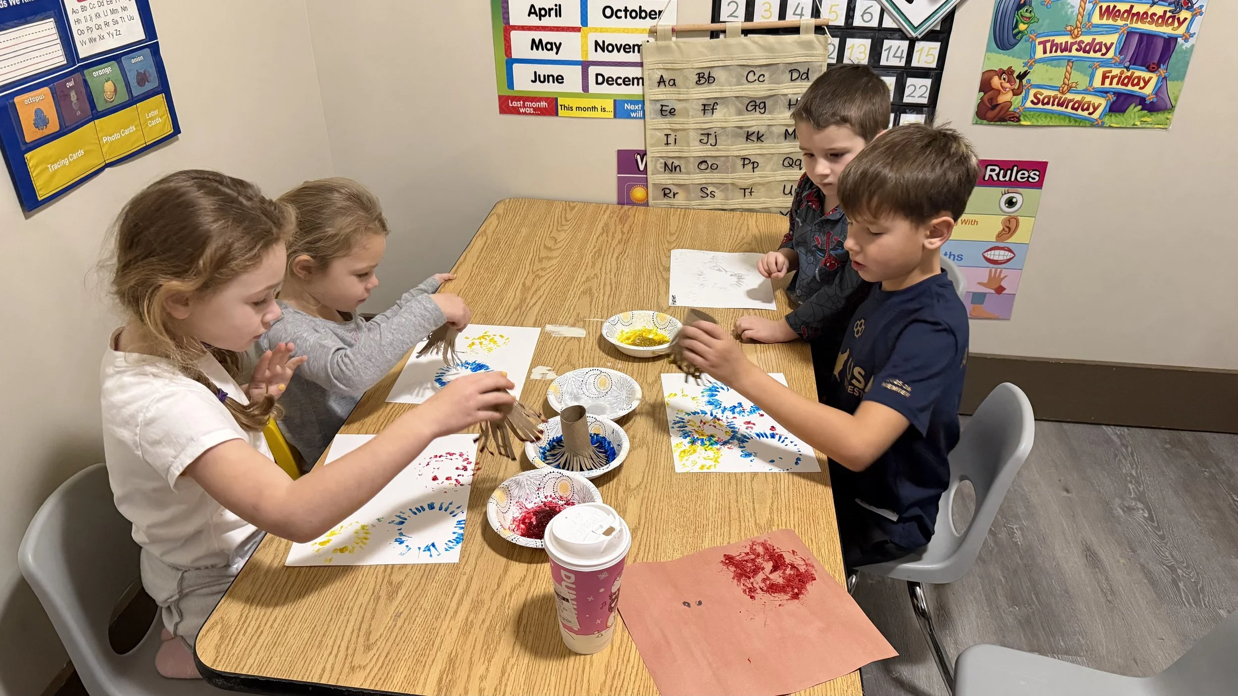 Four children sit at a table in a classroom, engaged in arts and crafts, painting and decorating paper with various colors and materials.