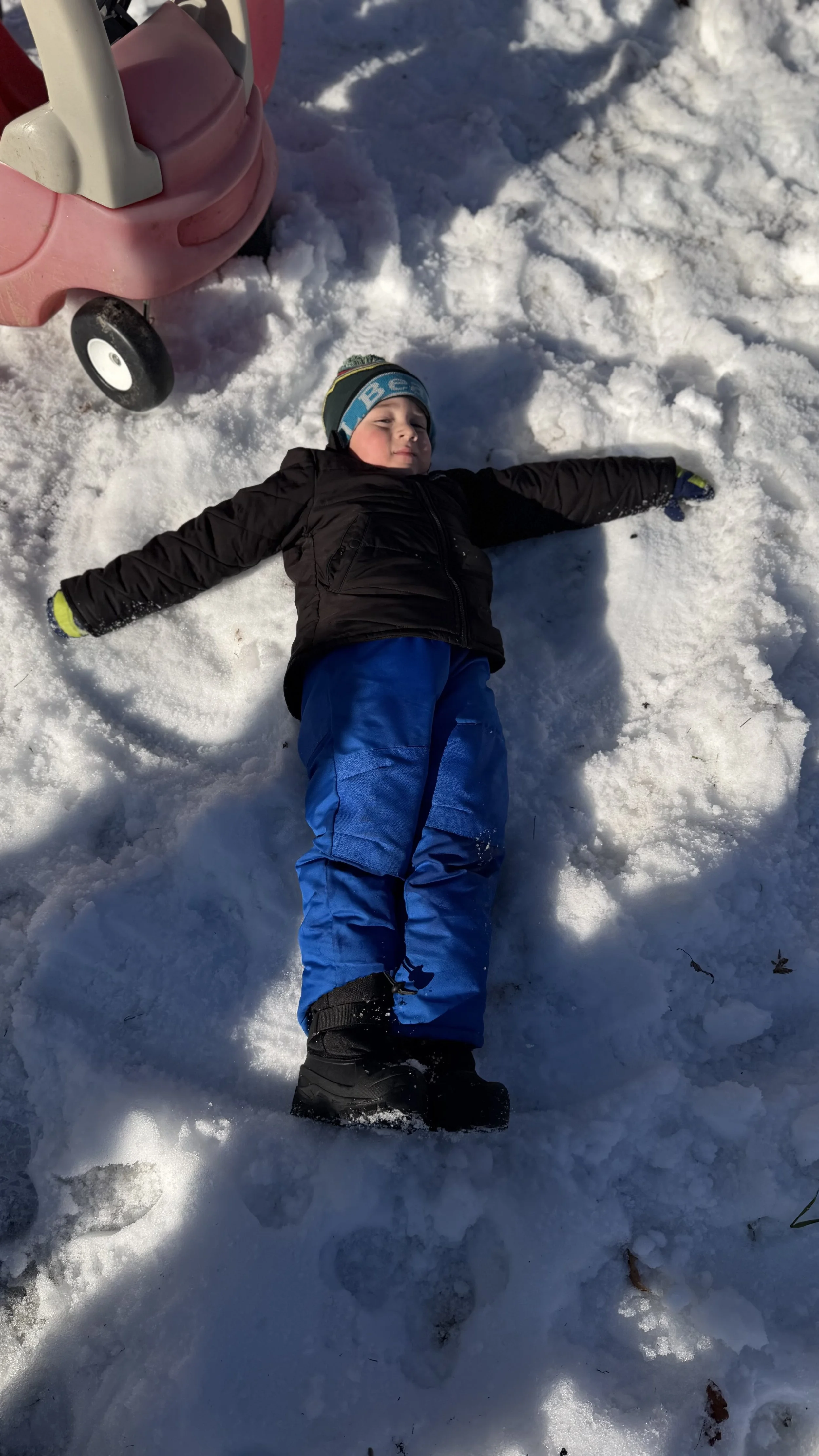 Child lying on snow making a snow angel during winter, with a pink stroller in the background.