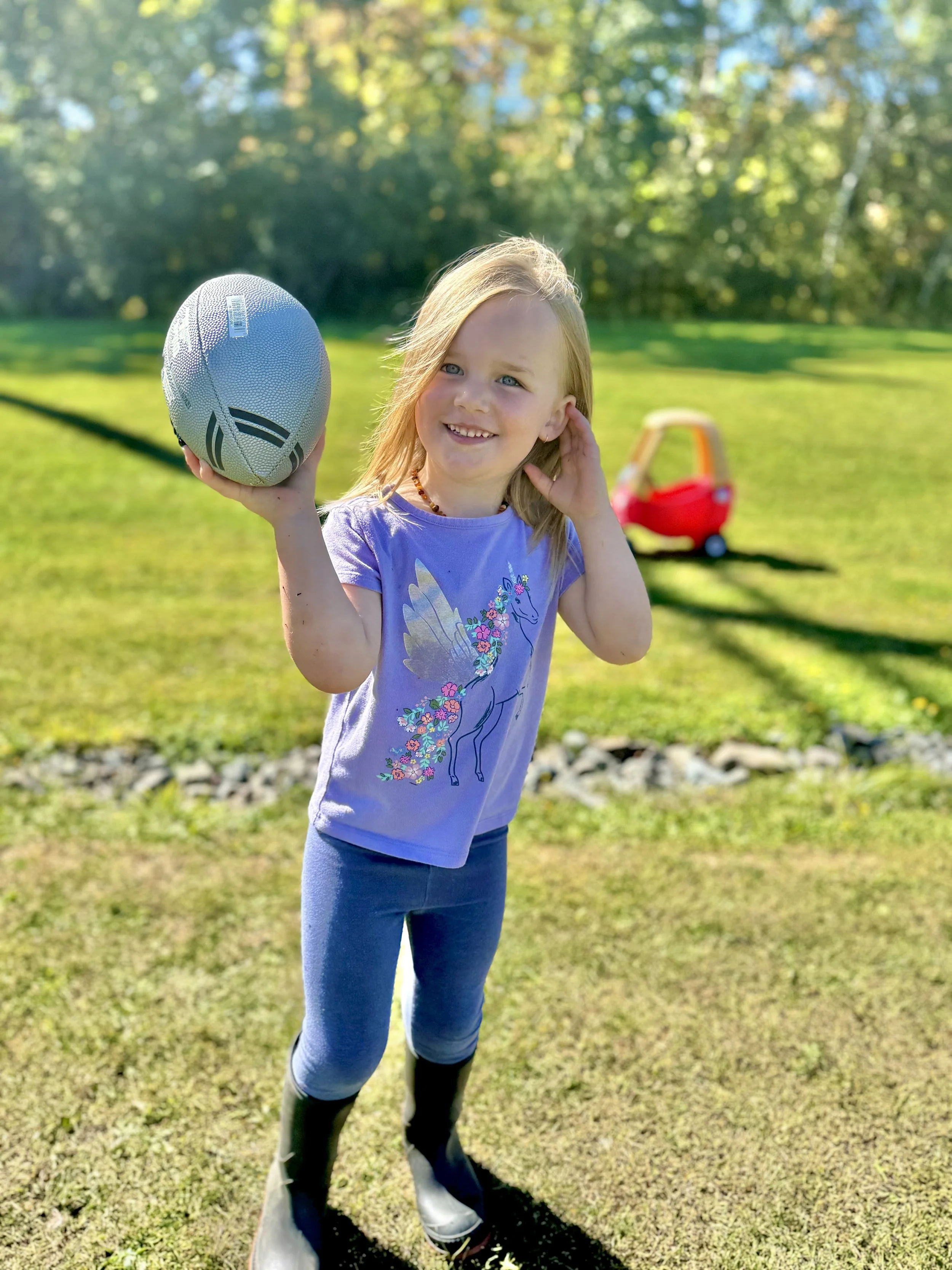 A young girl holding a silver football, standing outside on a grassy lawn with trees in the background, wearing a purple unicorn and fairy wings t-shirt, blue leggings, and black boots.
