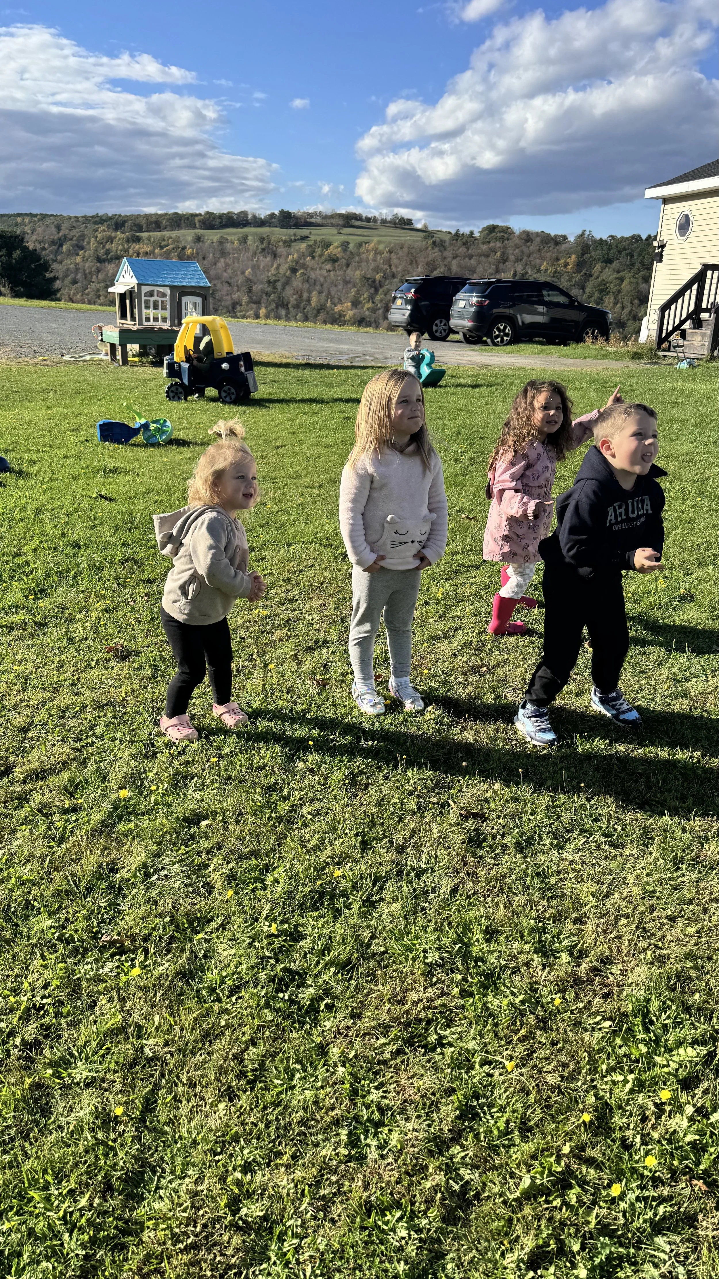 Four children outdoors on a grassy field, with some toys and a playhouse in the background, under a partly cloudy sky.