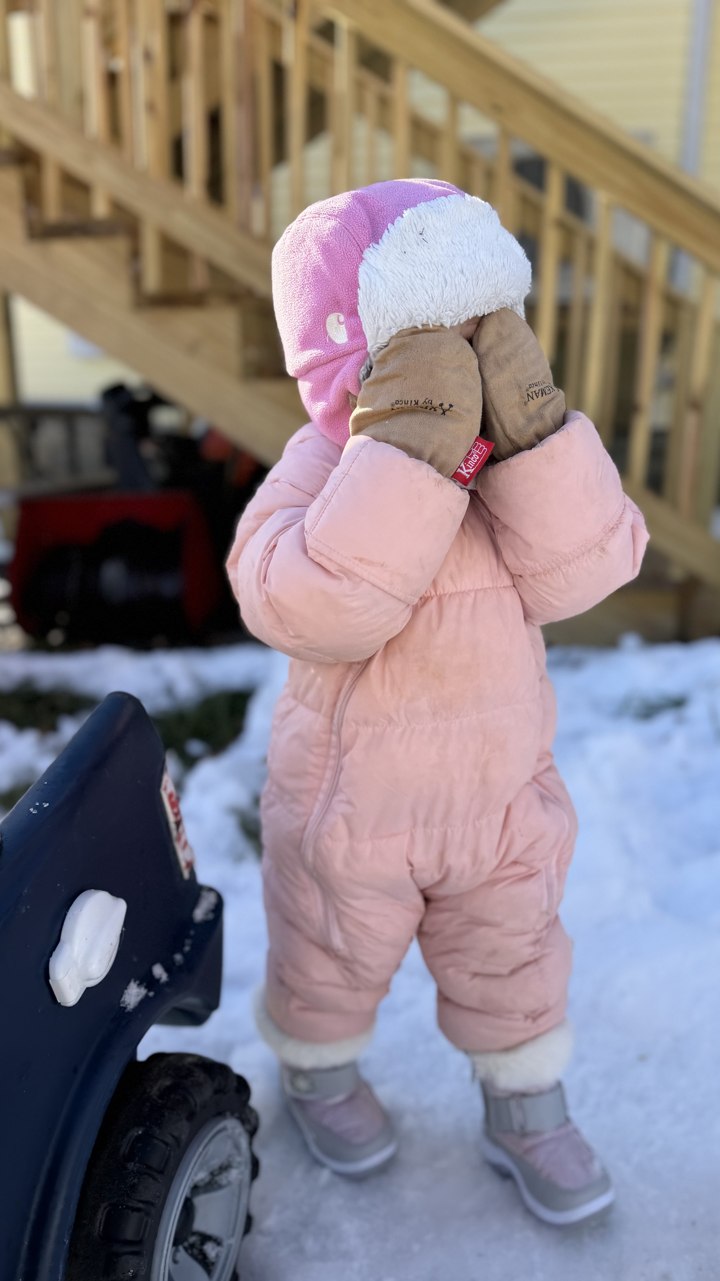 A young child dressed in a pink snowsuit, pink and white hat, and beige gloves, standing outdoors in the snow near a sled, with a wooden staircase in the background.
