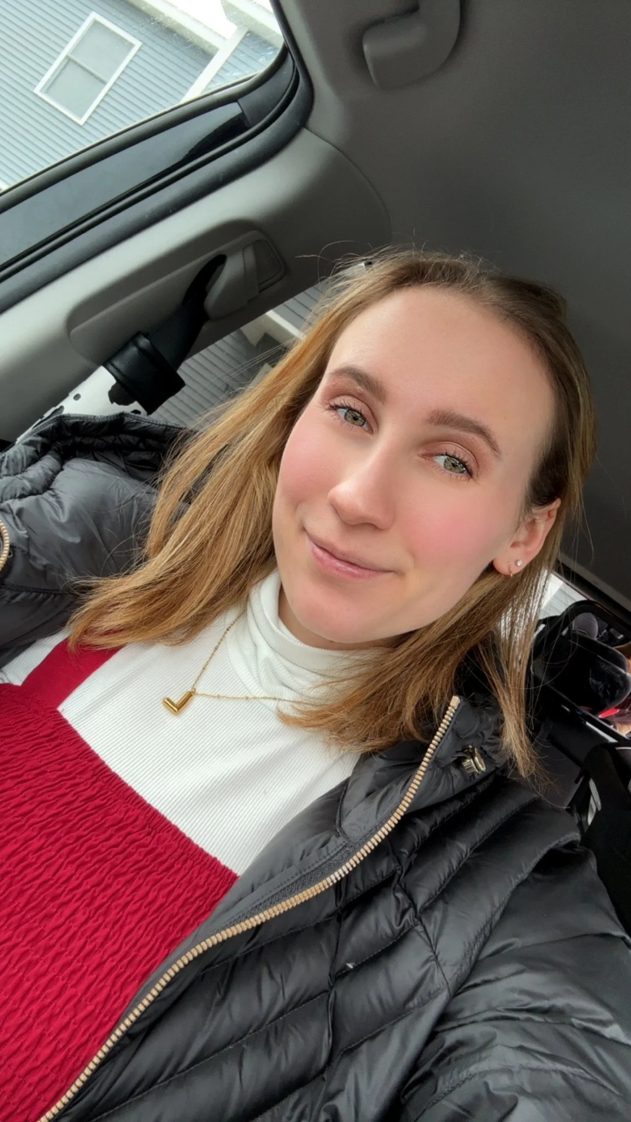 A young woman with light skin, auburn hair, and blue eyes taking a selfie inside a car. She is wearing a white turtleneck, a red textured top, a black puffer jacket, a gold necklace, and small earrings. The background shows a residential building with siding and windows.