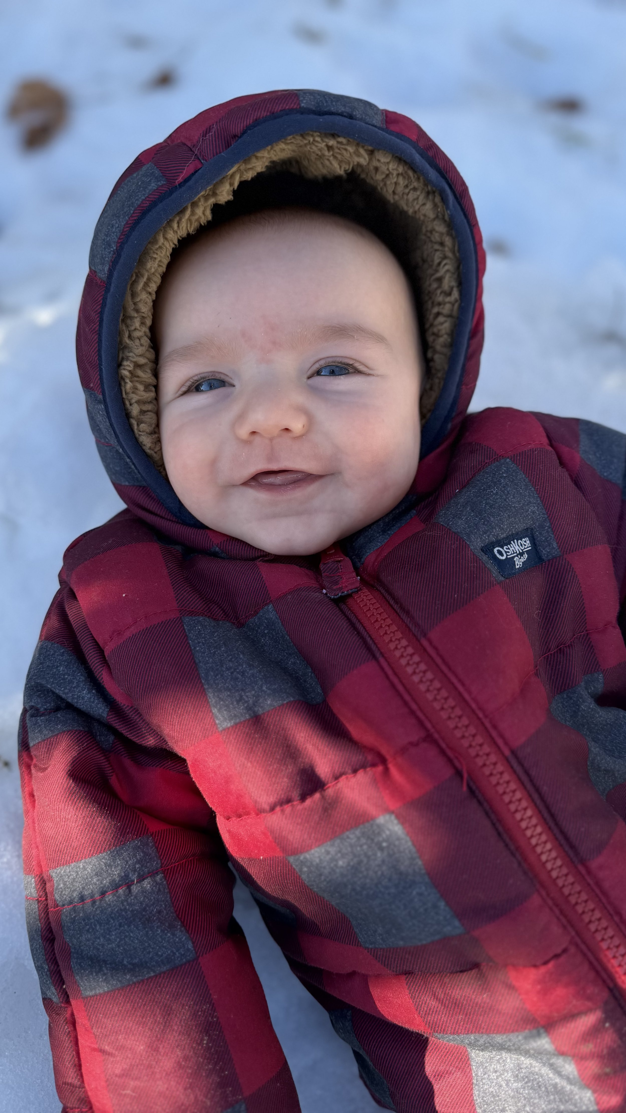 A smiling baby in a red and black plaid winter jacket and a warm hood, lying on snow.