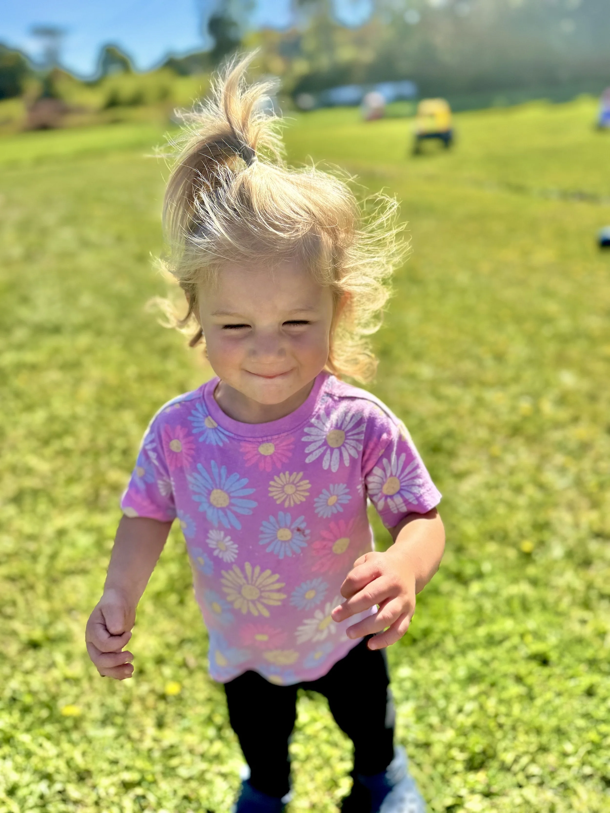 Young girl with blonde hair in a ponytail, wearing a pink shirt with daisies, running on a grassy field on a sunny day.