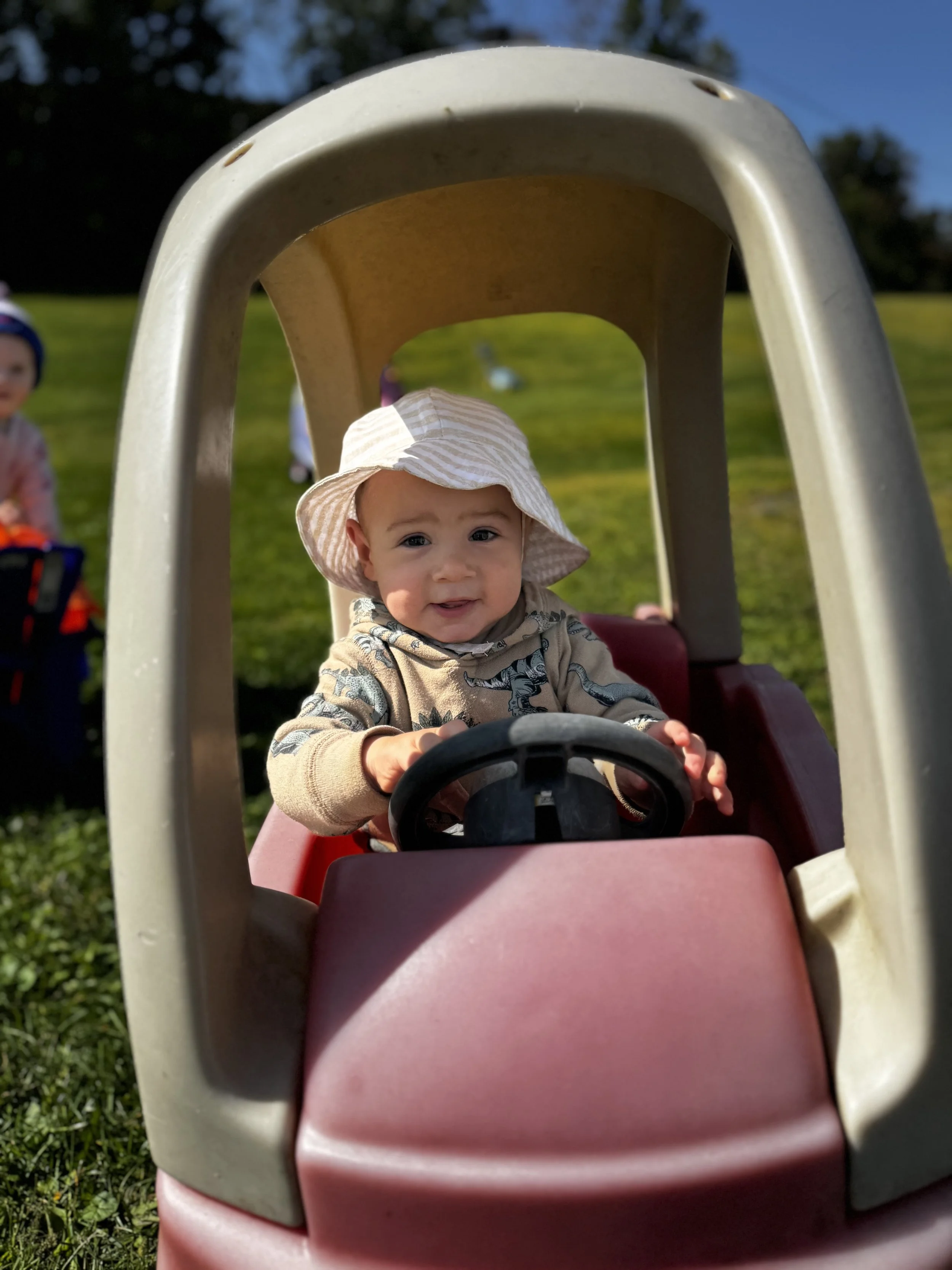 Young child wearing a striped sun hat and a dinosaur-printed hoodie sitting inside a red toy car outdoors on grass with other children in the background.