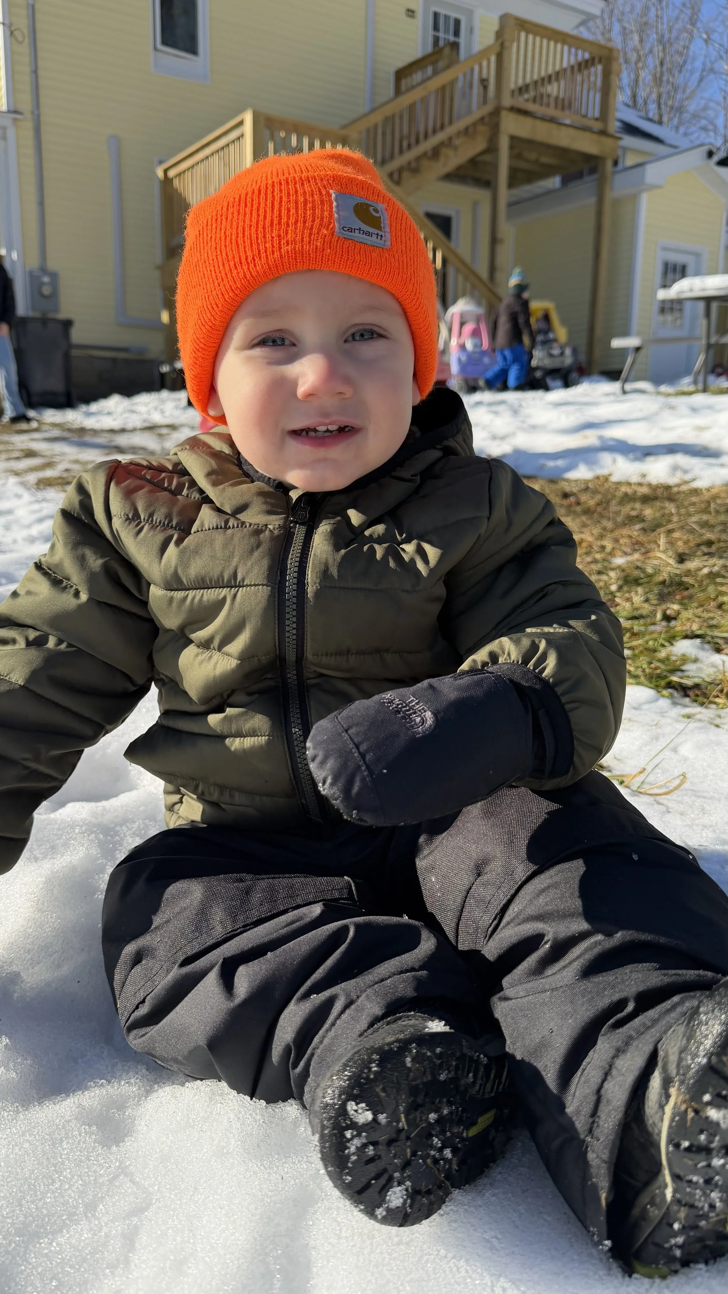 A young boy sitting on snow in a cold outdoor setting, wearing an orange Carhartt beanie, a green puffer jacket, black gloves, and snow boots, with a house and other people in the background.