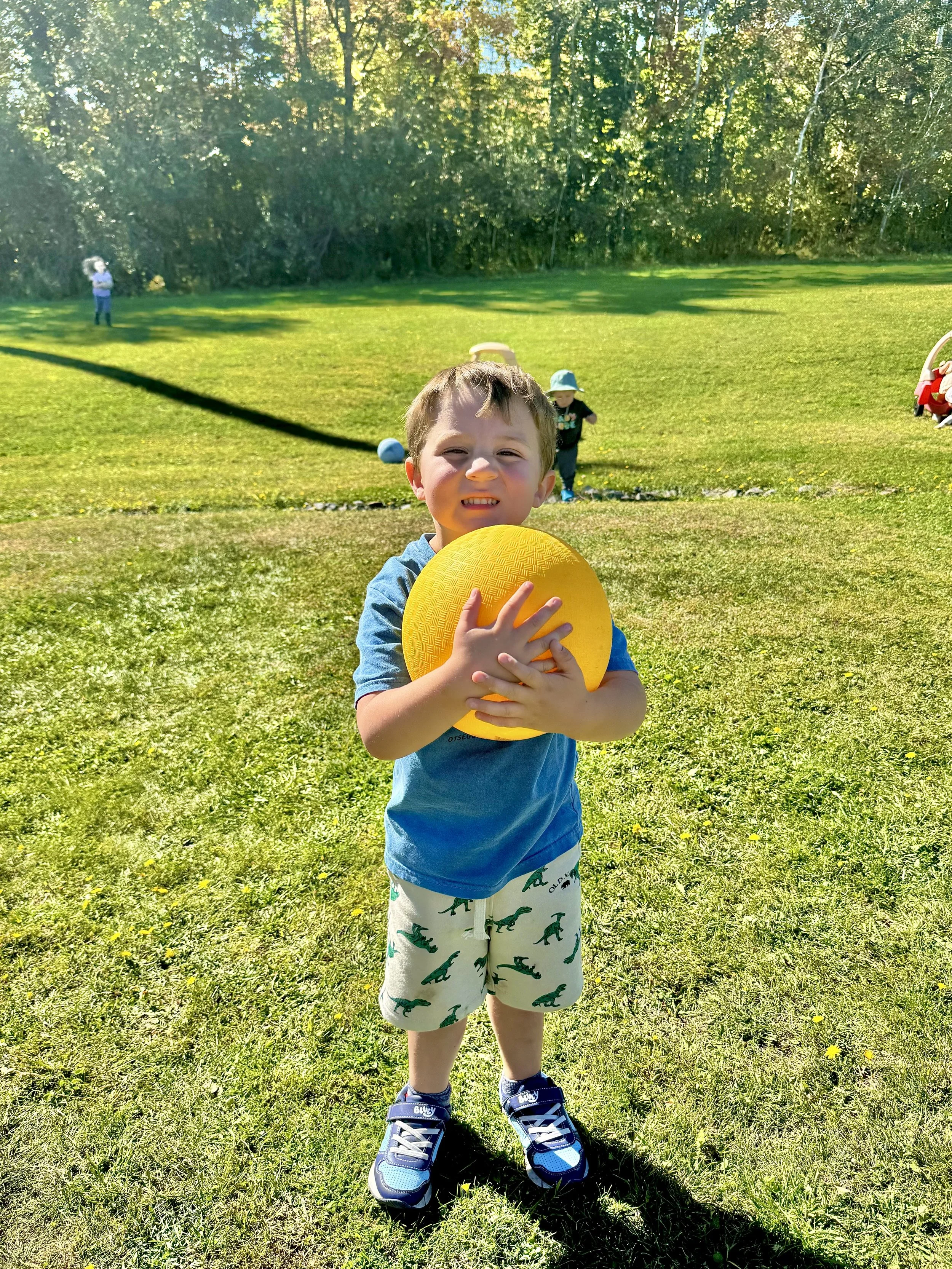 A young boy holding a yellow ball, outdoors on a sunny day, with other children playing in the background on a grassy field and surrounded by trees.