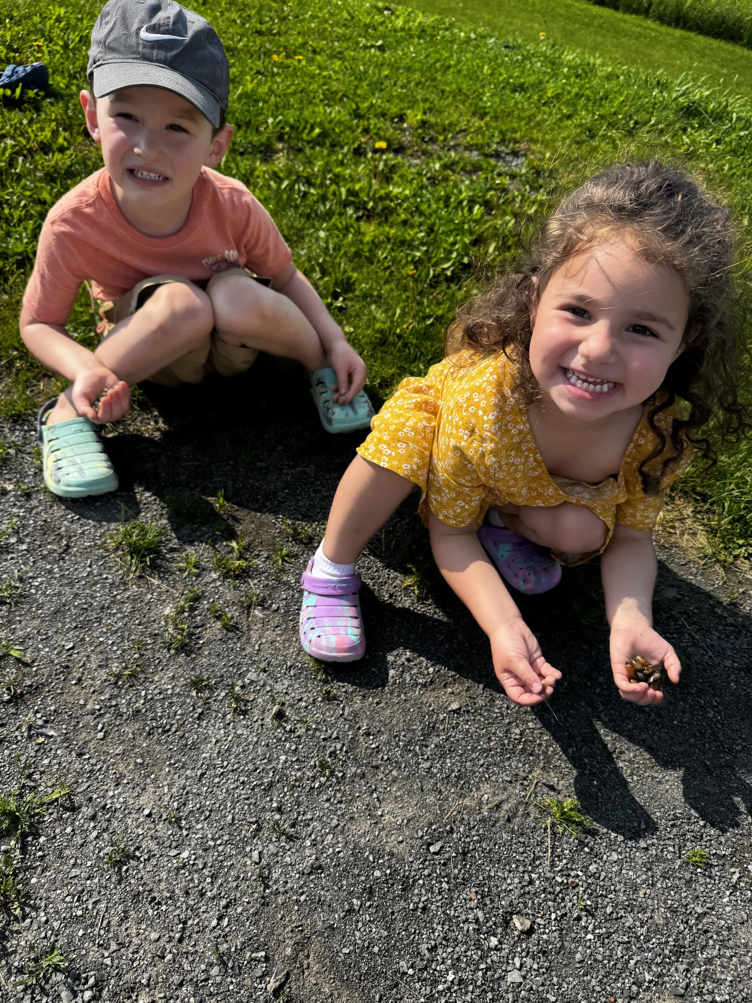 A young boy and girl are crouching on a gravel path outdoors, smiling at the camera. The girl is holding a small turtle in her hand. The background is green grass and some plants.