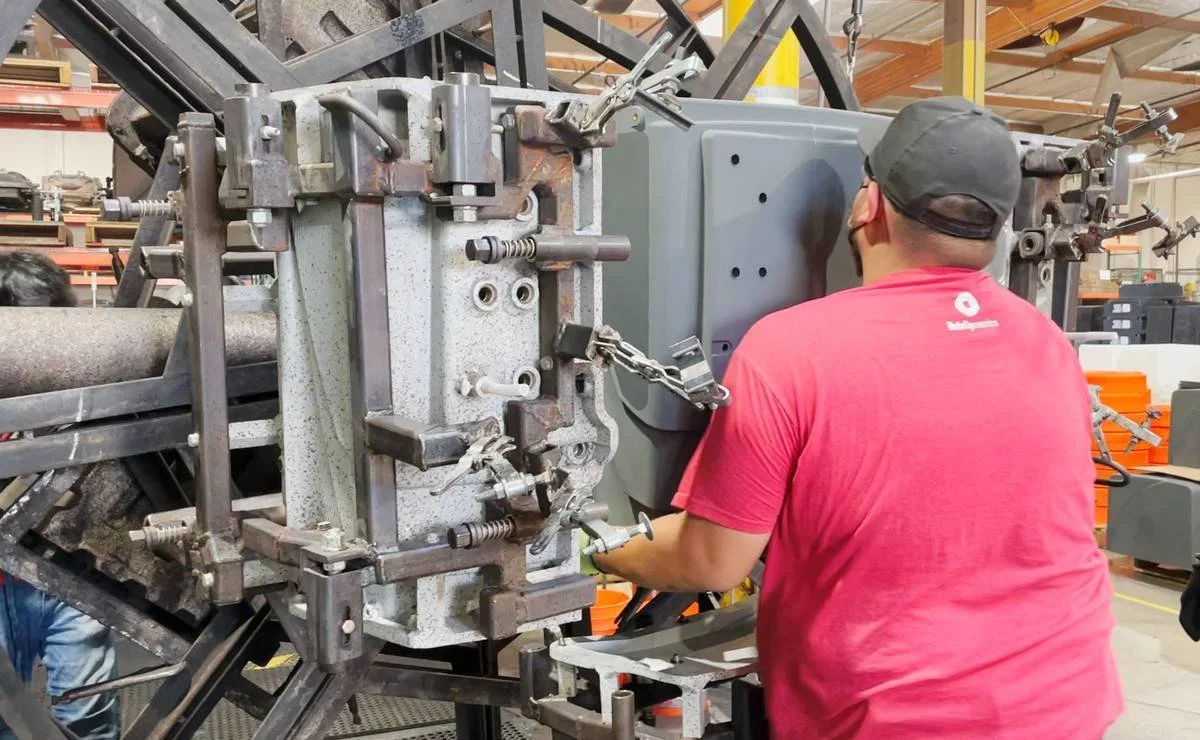 A man in a red shirt and black cap working on rotational mold industrial machinery in a manufacturing factory.
