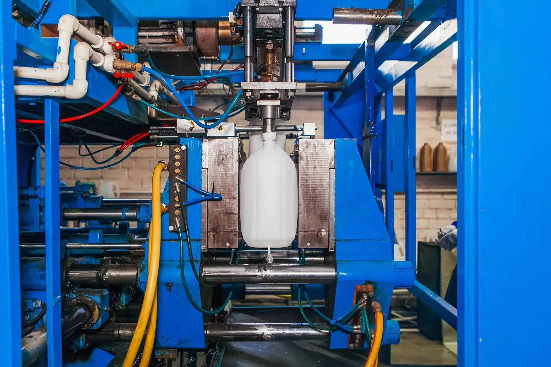 A machine with a white plastic bottle secured in the center, surrounded by metallic and blue components, with various pipes and wires connected, in an industrial setting.