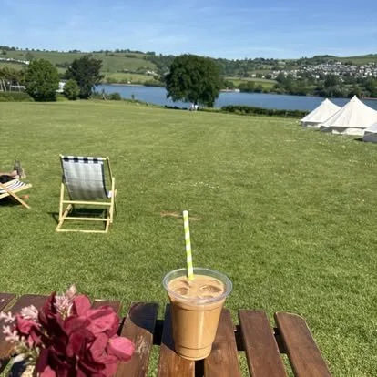 A drinks on a wooden table with pink flowers in the foreground, overlooking a grassy park with lounge chairs, tents, a lake, and trees in the background on a sunny day.