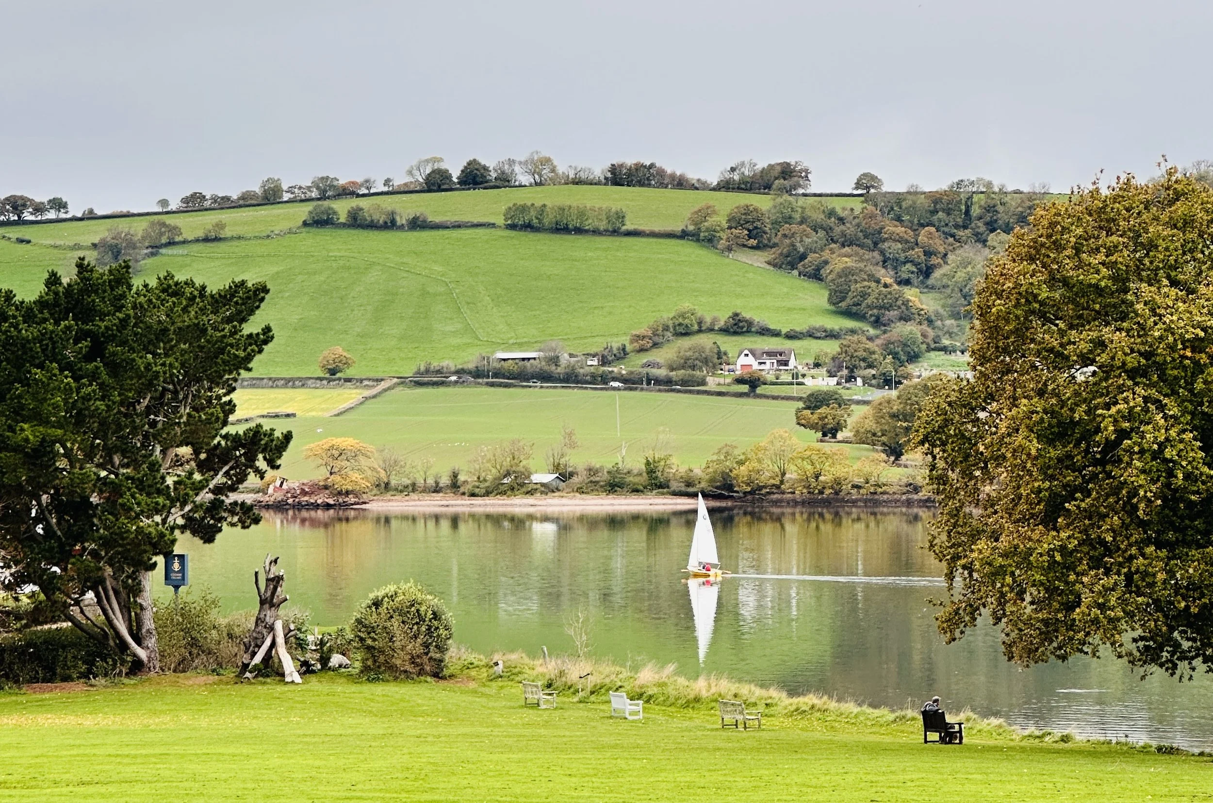 A scene of a lake with a sailboat, surrounded by green grass, trees, and rolling hills in the background.