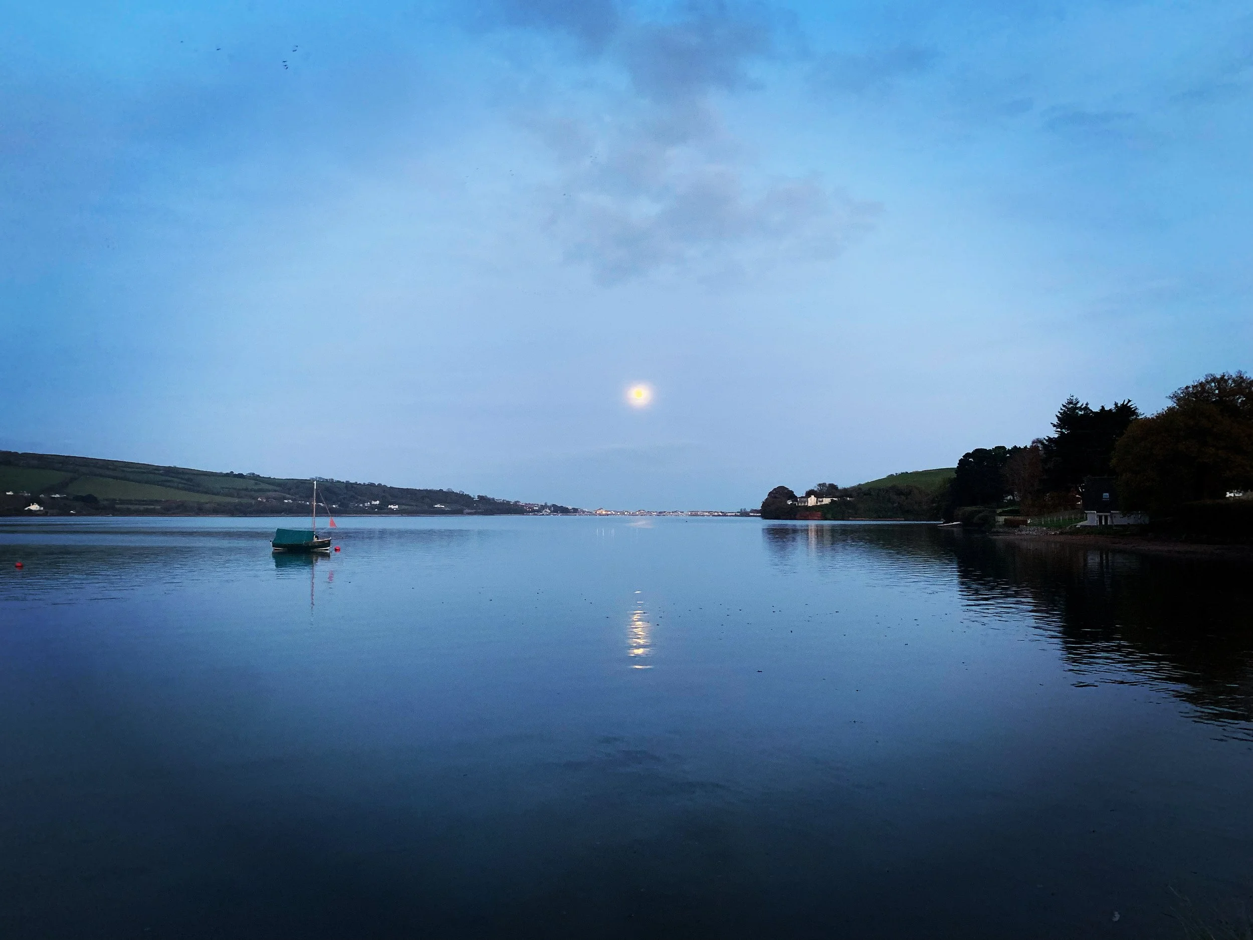 A calm body of water reflects the sky with a bright moon. A small boat is anchored on the water, and a few houses and trees line the shoreline on either side.