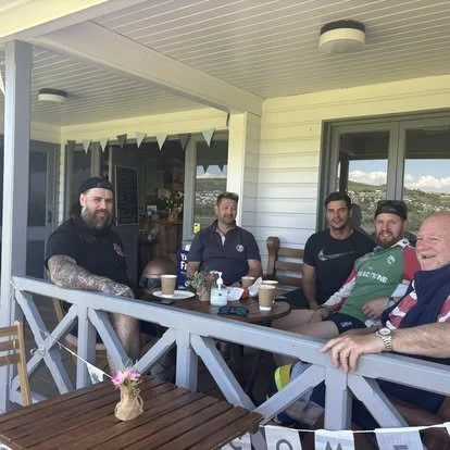 Five happy men drinking coffee and  sitting on a porch at a table with coffee cups, a small pot with pink flowers, and a sunny outdoor view in the background.