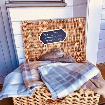 A wicker bench with a blackboard sign which reads 'Feel Free to Borrow & Use a Blanket'. There are several folded blankets on the bench.