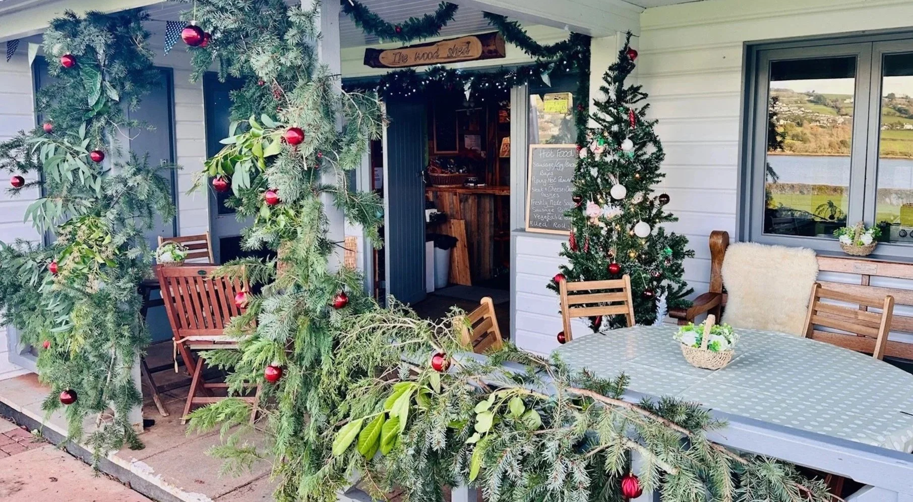Outdoor patio decorated for Christmas with a Christmas tree and a festive arch made of greenery and red ornaments, wooden chairs, and a table with a green polka dot tablecloth, a basket of flowers, and a cozy chair with a blanket, overlooking a sceni