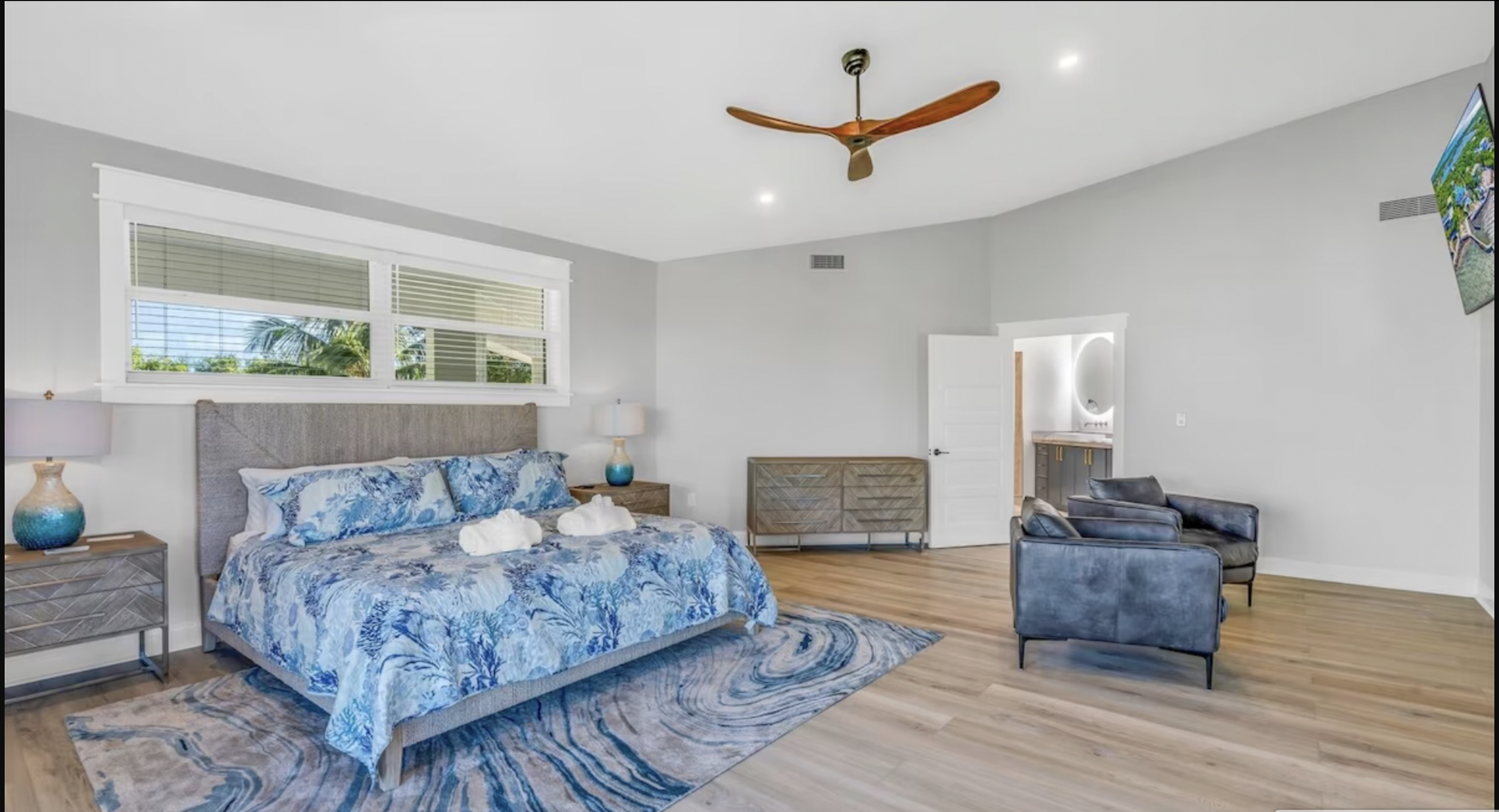 Luxurious bedroom with ocean view, featuring a large bed, cushioned chairs, flat-screen TV, fireplace, and a sunset visible through large windows.