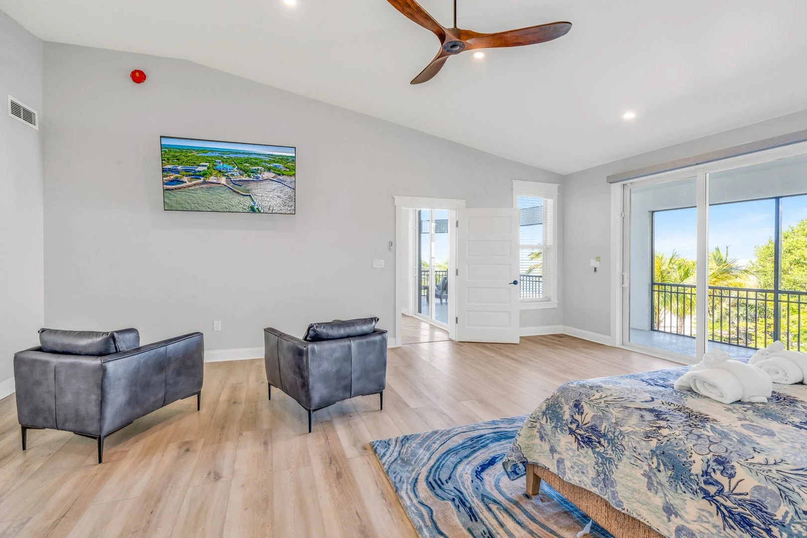 Luxurious bedroom with ocean view, featuring a large bed, cushioned chairs, flat-screen TV, fireplace, and a sunset visible through large windows.
