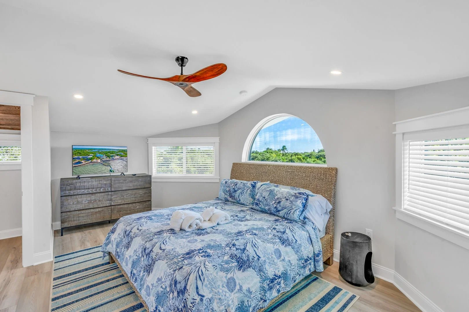Modern bedroom with large bed, decorative pillows, bedside table with lamp, wall-mounted TV, and seating area near a window.
