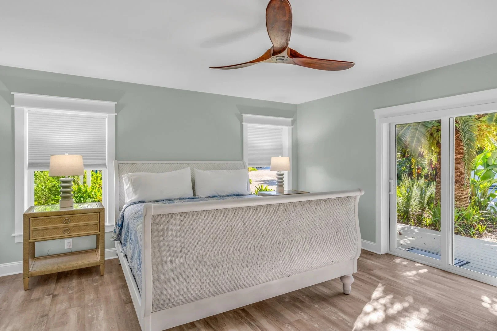 A bedroom with a wooden bed, white bedding, pillows, a nightstand with a lamp and clock, a dresser with a TV above, a ceiling fan, and decorative mirrors on the beige walls.