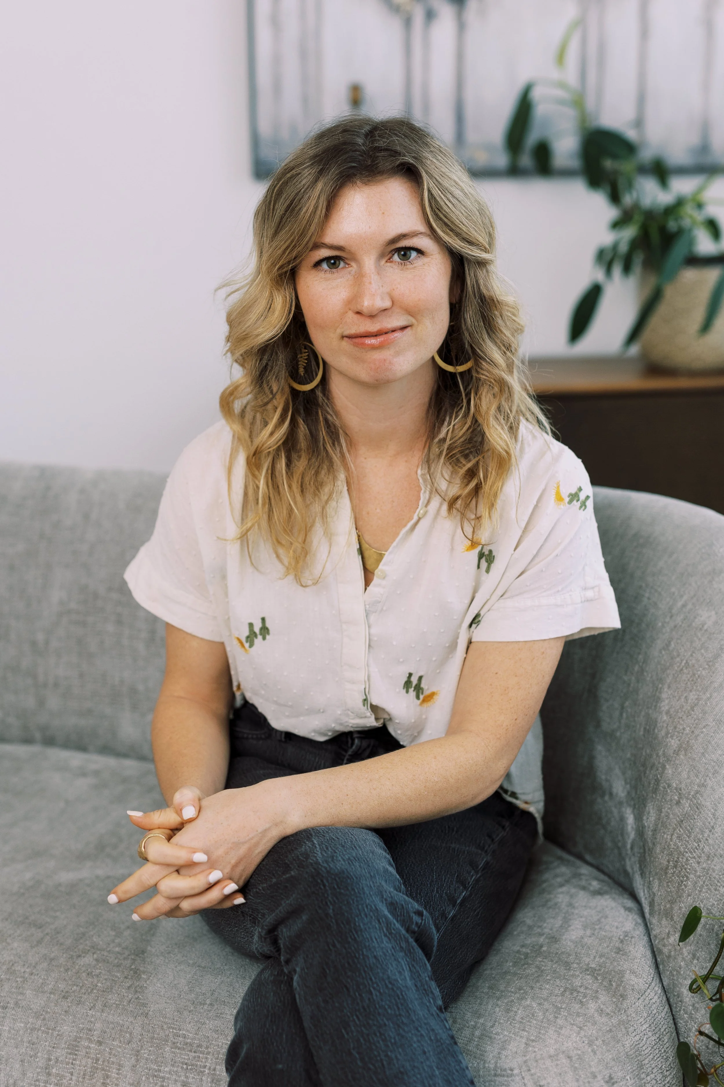 A woman with wavy blonde hair and blue eyes sitting on a gray couch, wearing a white shirt with small cactus embroidery, gold hoop earrings, and dark jeans, with green plants and artwork in the background.