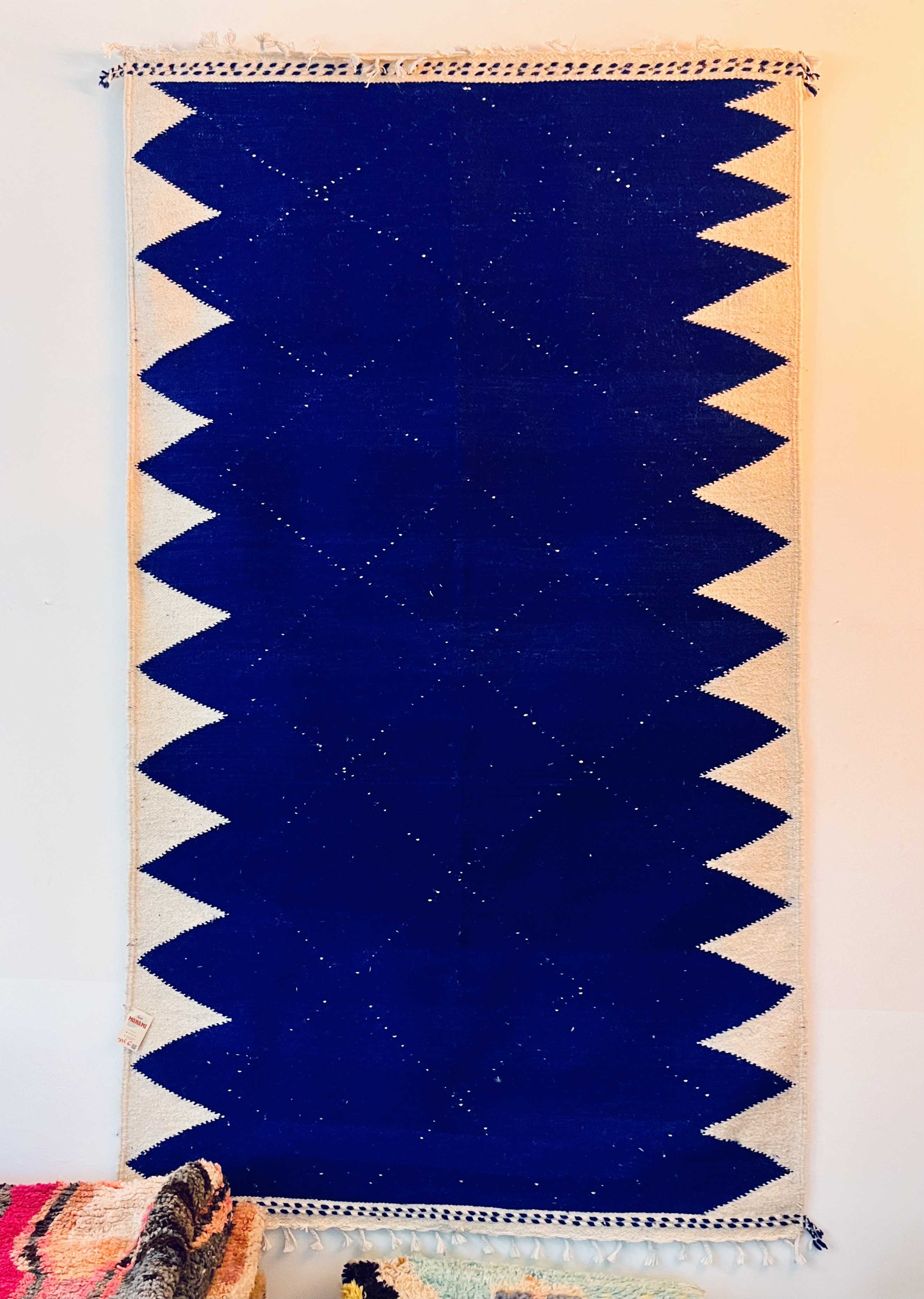 A vertically oriented woven rug with a dark blue central section decorated with white star-like patterns. The border features zigzag edges in beige and dark blue.